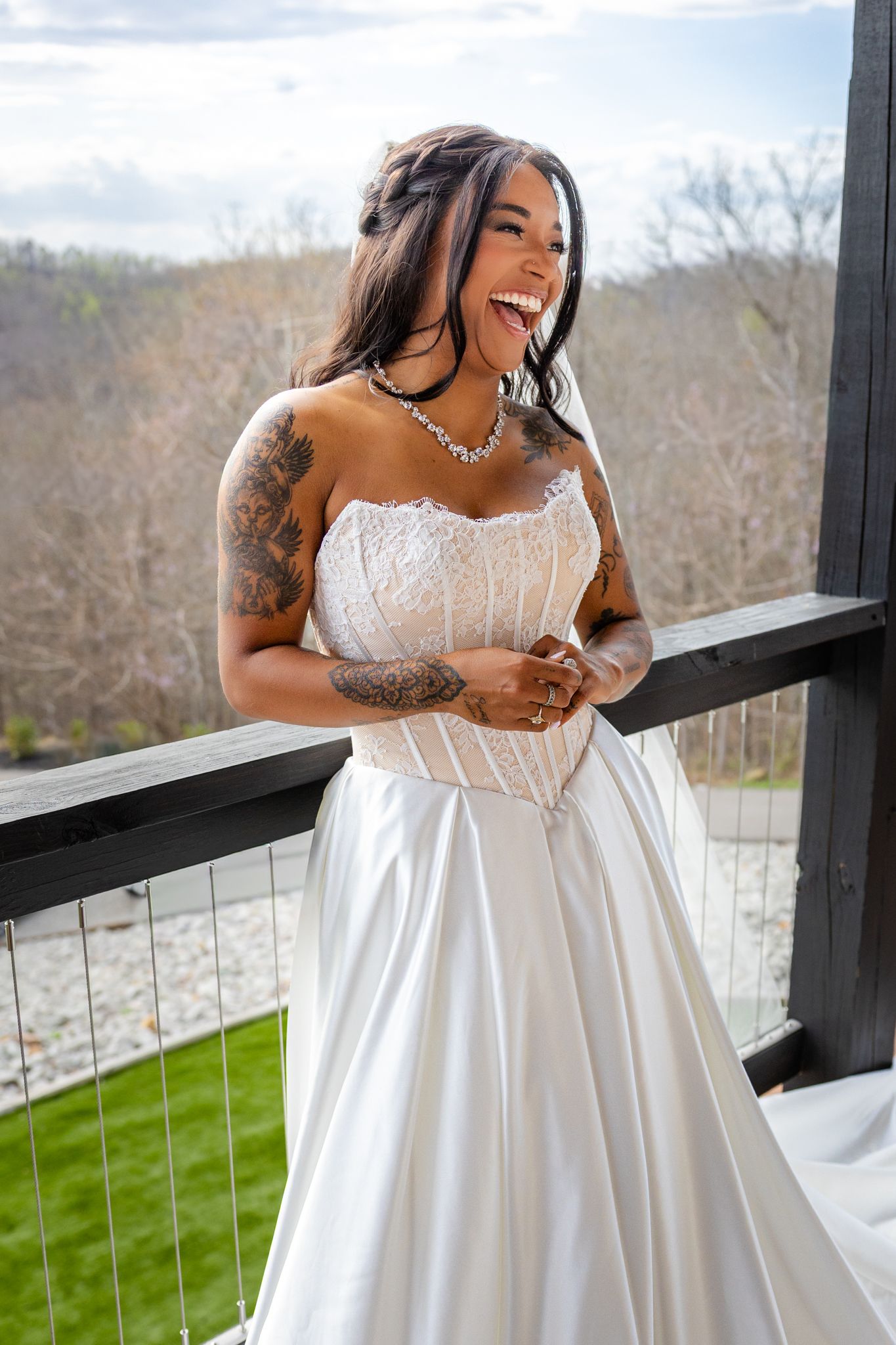 Laughing bride in strapless lace corset wedding gown with tattoos poses on outdoor balcony.