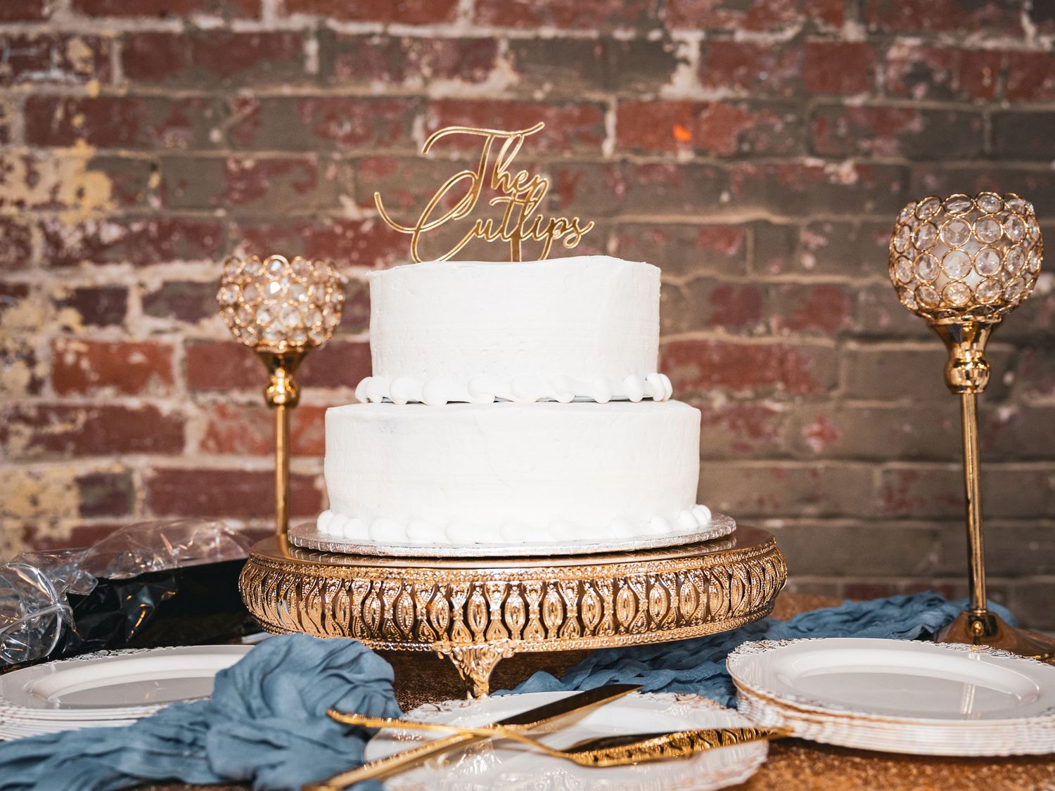Elegant two-tier white wedding cake with gold script topper displayed on ornate gold cake stand.