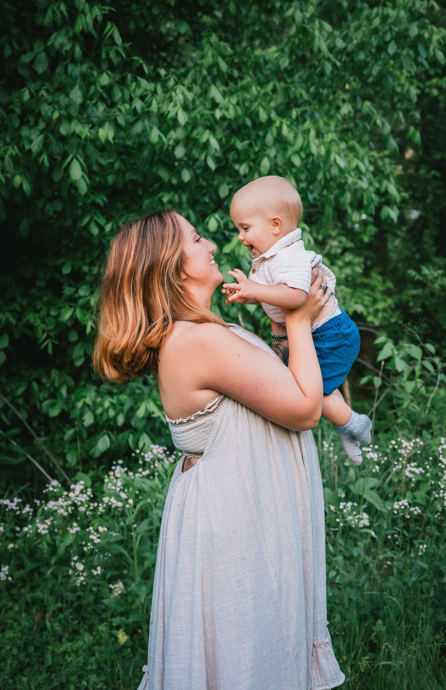 Mother in white dress lifting laughing baby boy in lush green garden setting.
