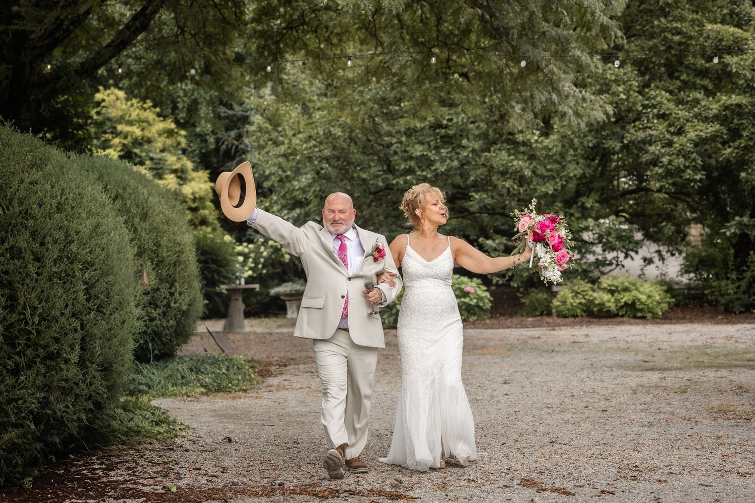 Joyful newlyweds celebrating outdoors, groom raising hat in air, bride holding pink bouquet, walking through lush garden path.