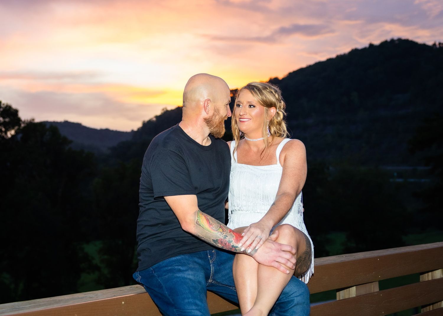 Couple sharing a romantic moment on a deck at sunset with mountain and colorful sky backdrop.