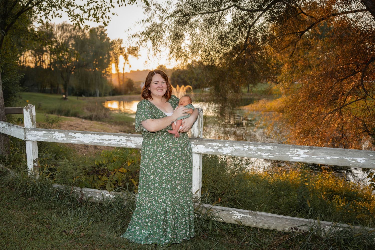 Woman in green floral dress holds newborn baby by white fence at golden hour sunset near pond.