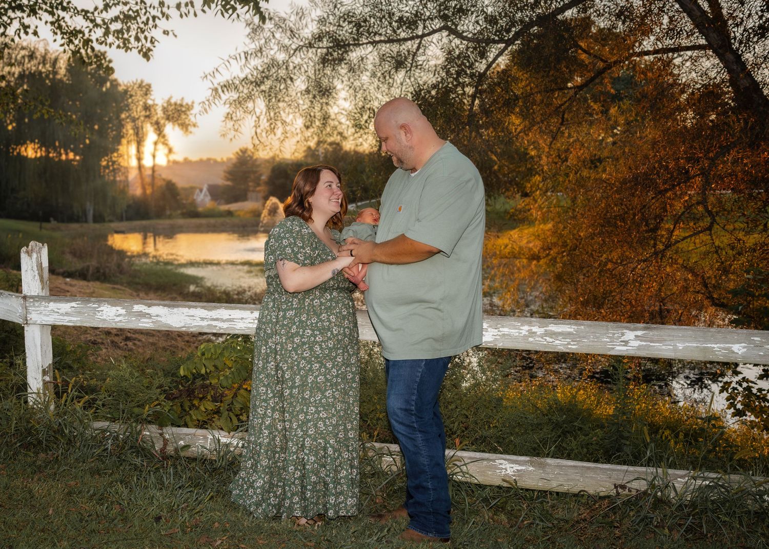 Couple dancing by white fence at golden sunset with autumn foliage and pond in background.