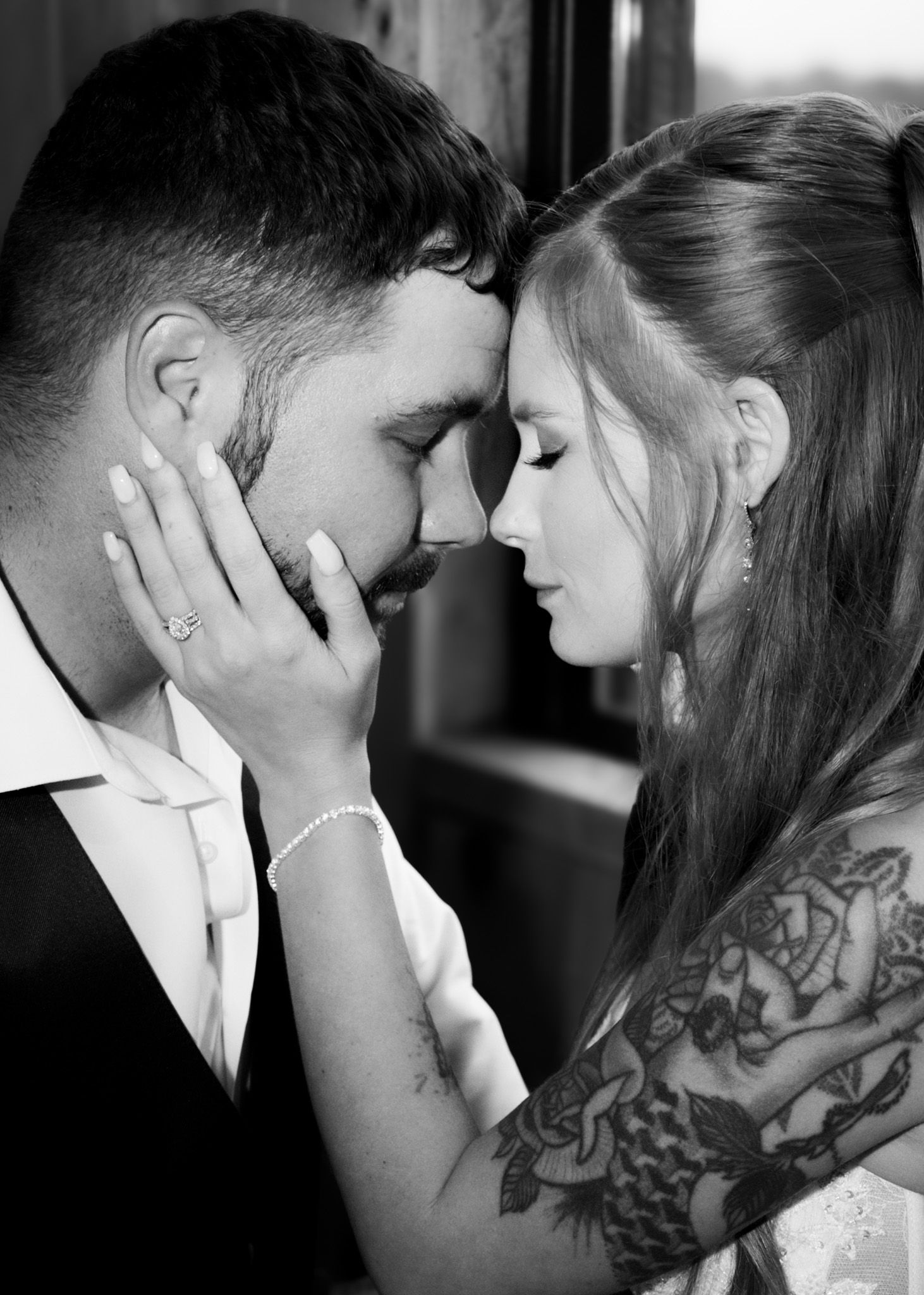 Black and white photo of a tattooed bride gently cradling grooms face in an intimate moment.