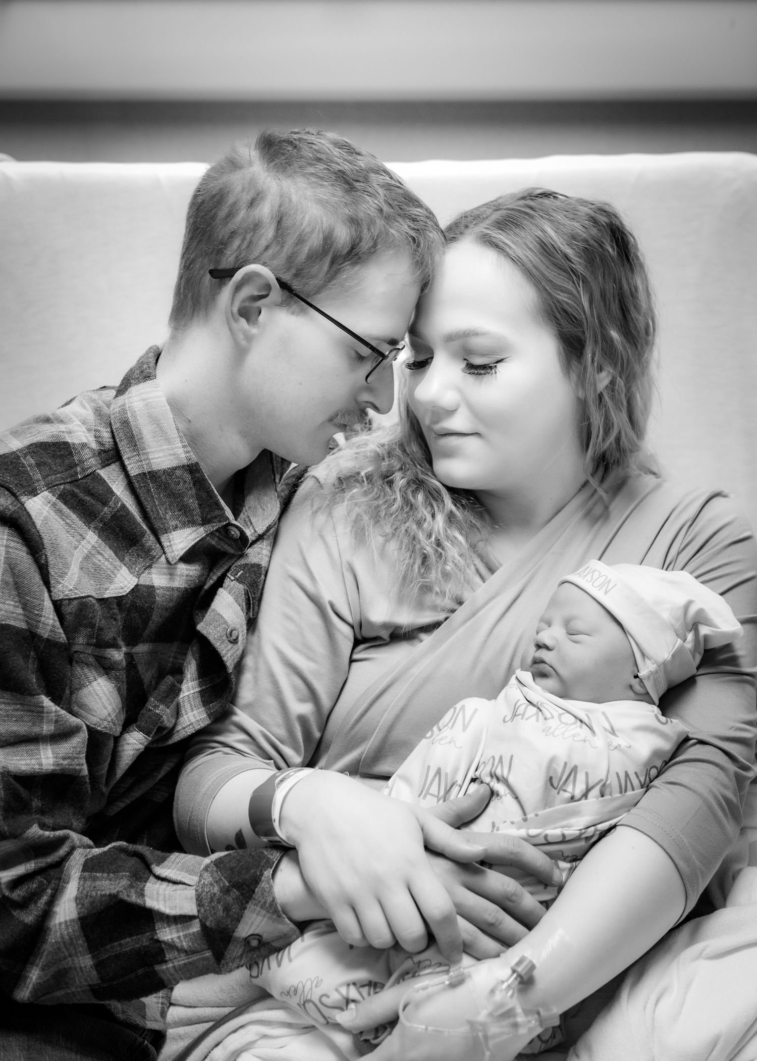 New parents gazing lovingly at their sleeping newborn baby in hospital room, tender black and white portrait.