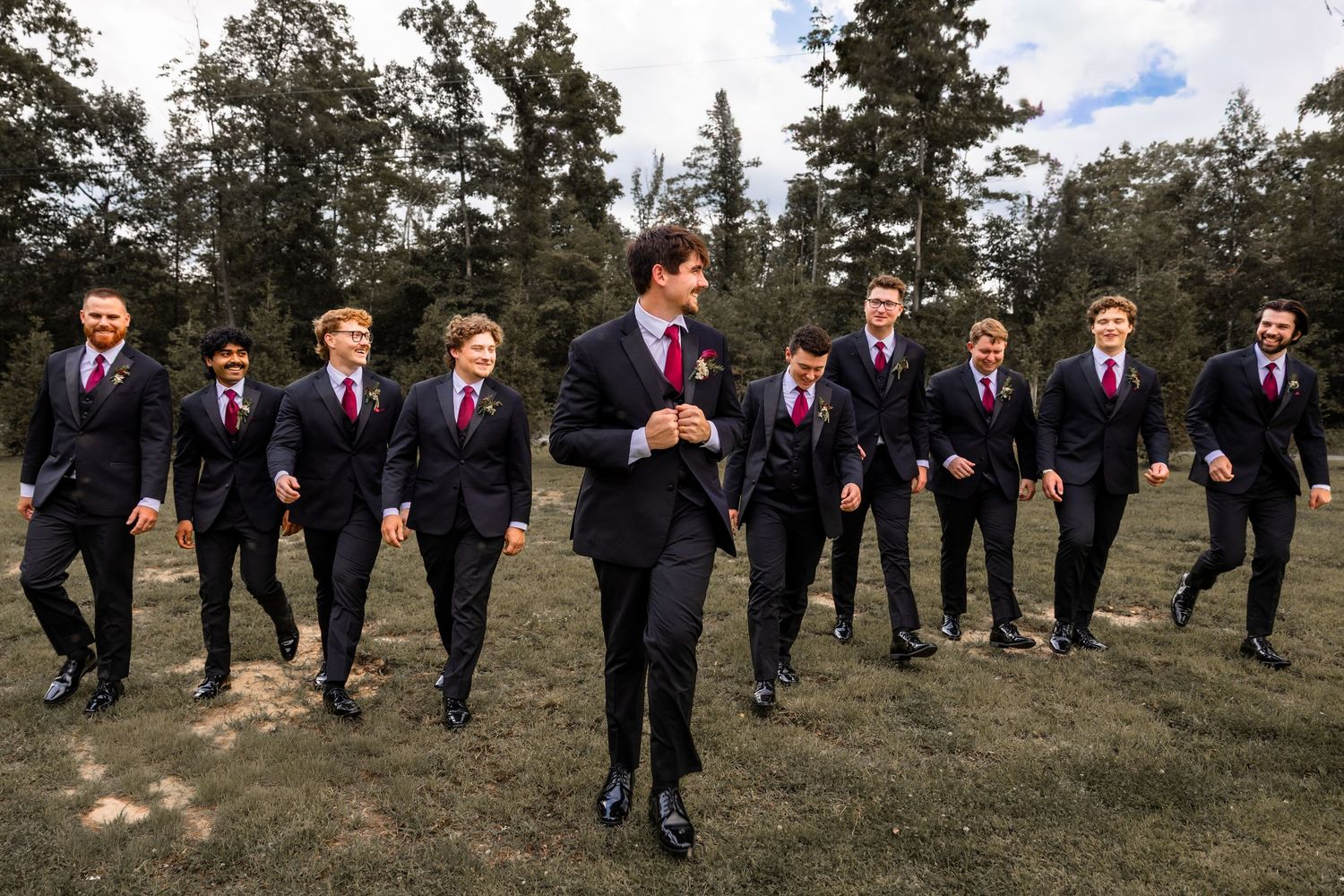 Groom and groomsmen in black suits with burgundy ties walking outdoors on grass with trees in background.