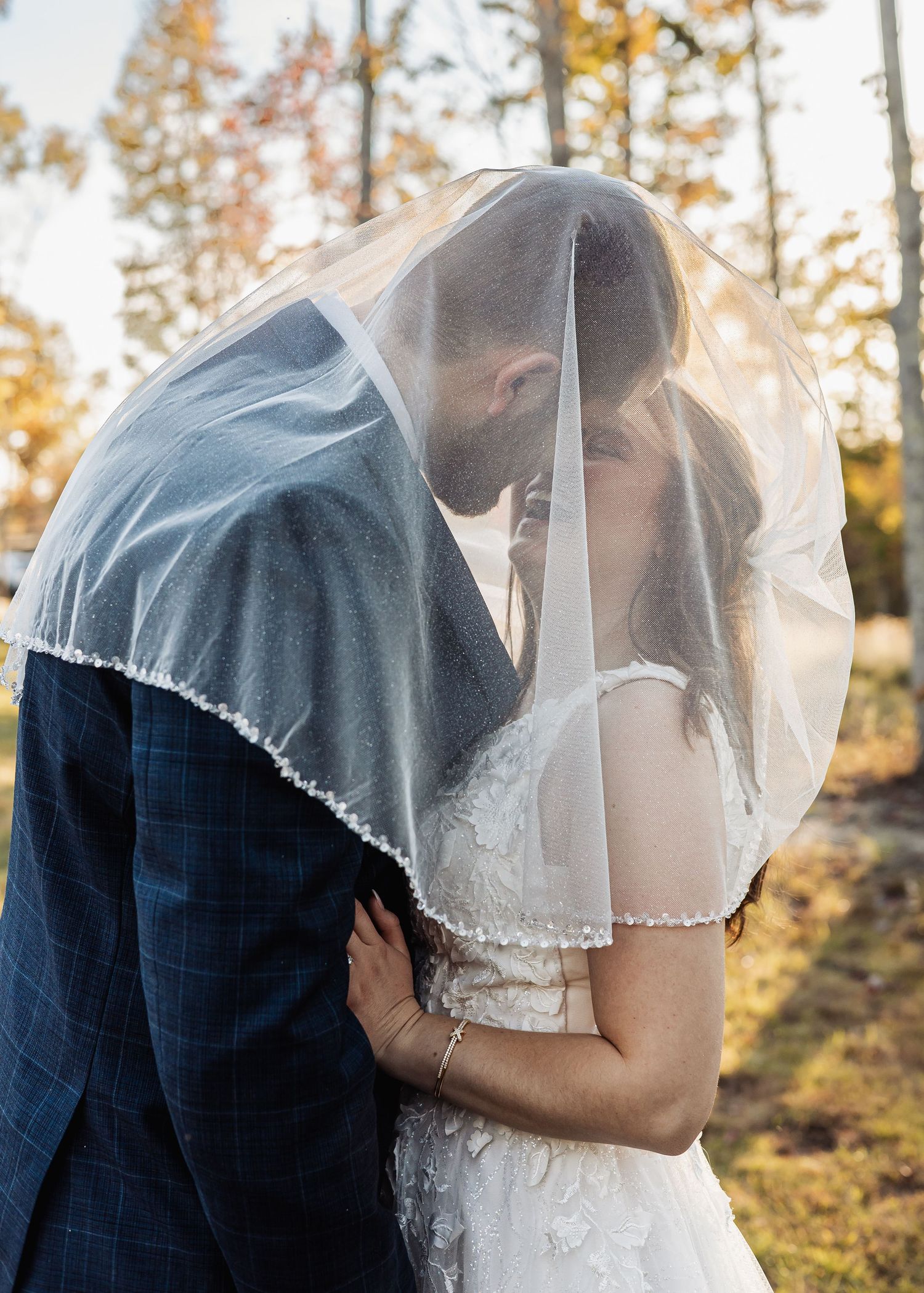 Bride and groom sharing a romantic kiss under a flowing white wedding veil outdoors in autumn.