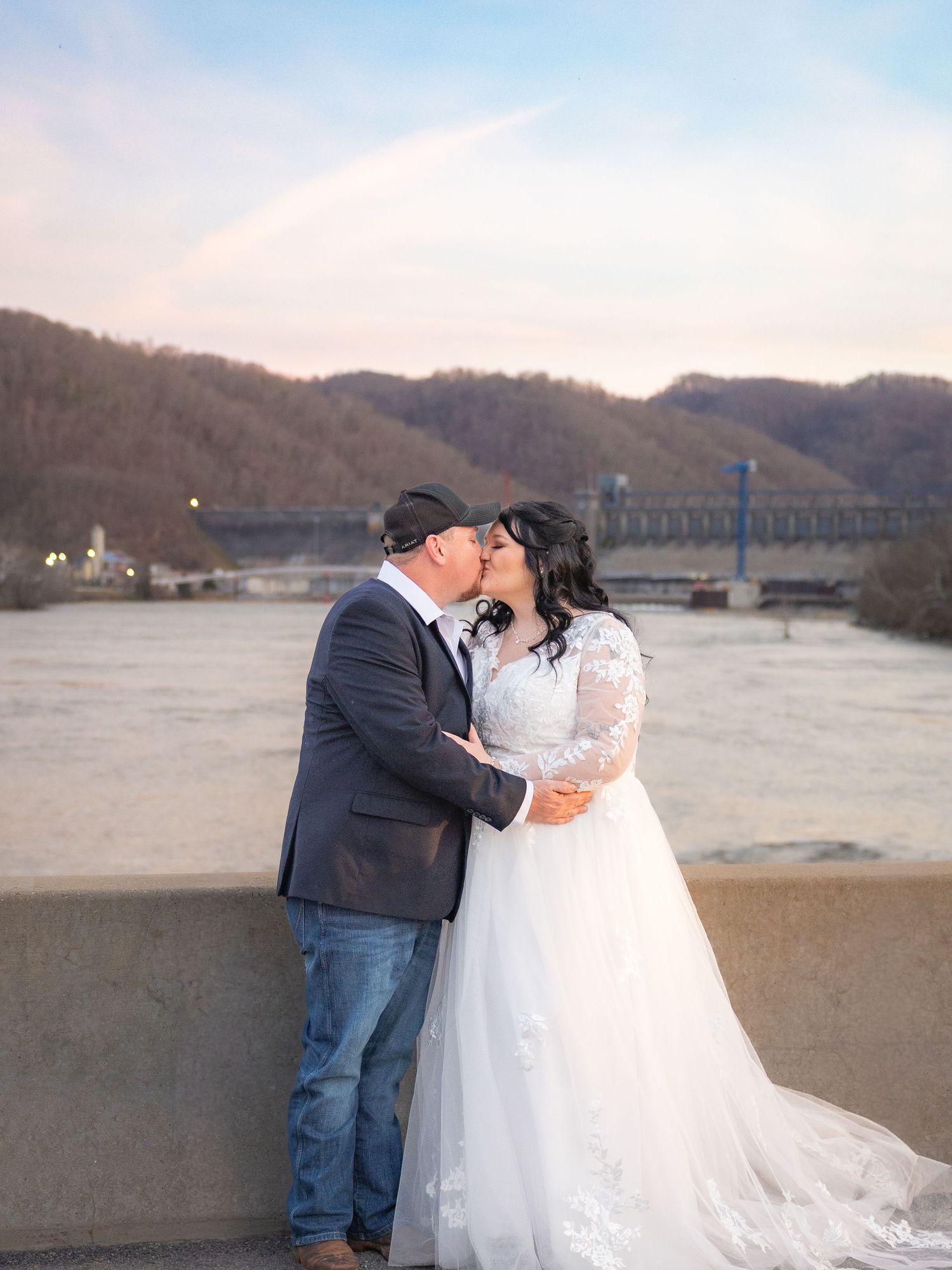 Bride and groom share a kiss outdoors with a river and bridge in background.