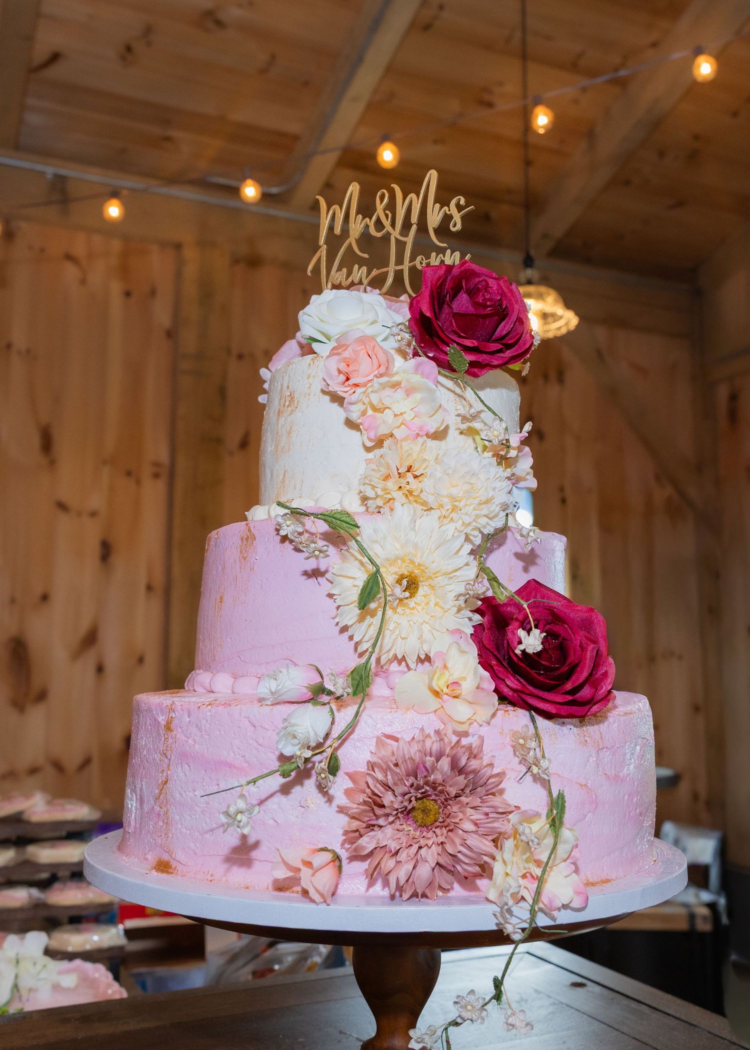 Four-tier pink and white wedding cake with fresh floral decorations and gold Mr & Mrs topper displayed in rustic barn venue.