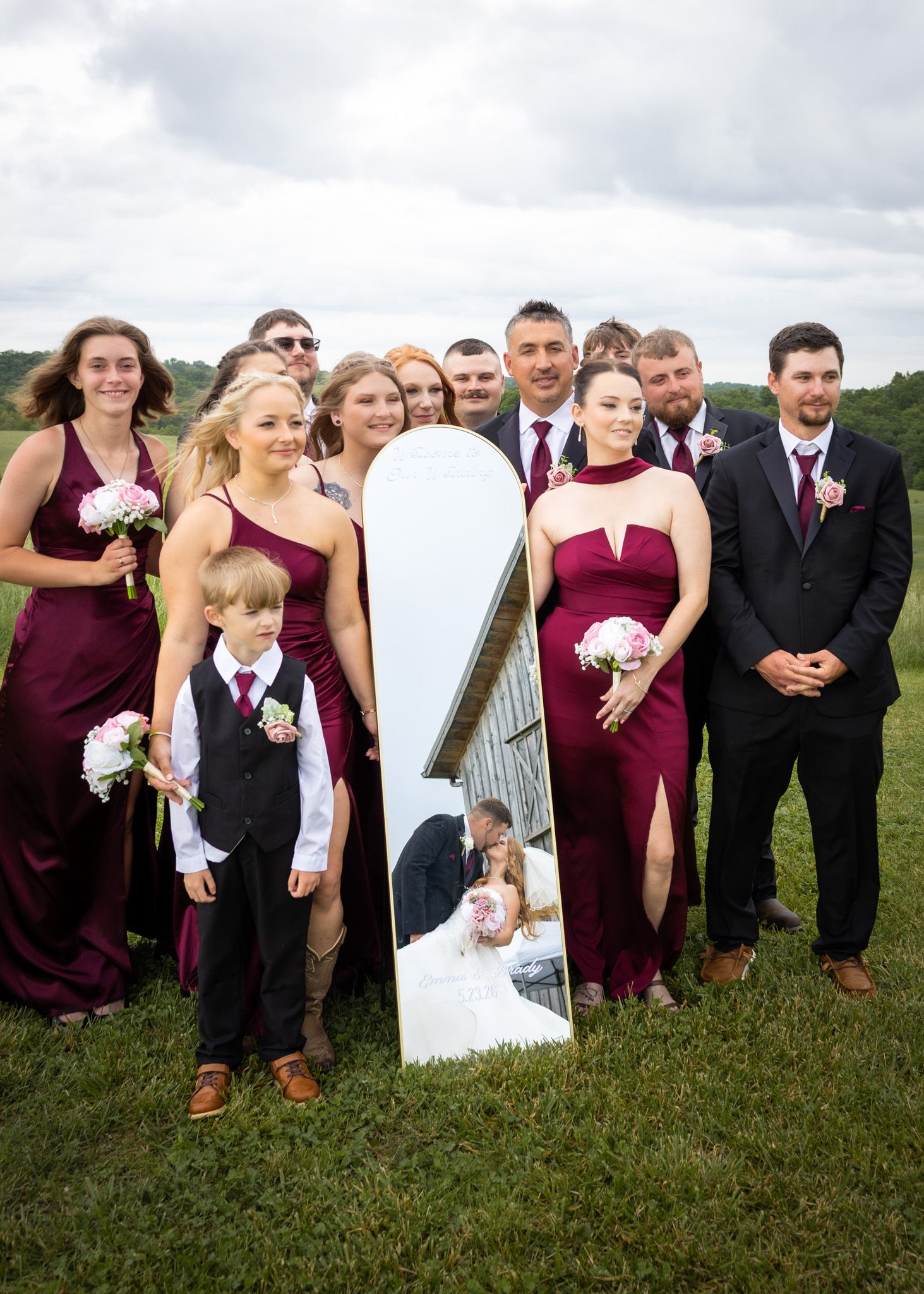 A wedding party in burgundy and black poses outdoors with a mirror reflecting the bride and groom kissing.