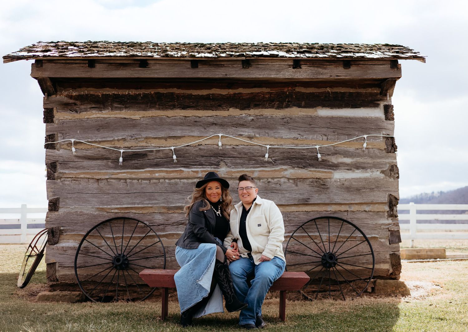 Smiling couple sitting on a bench in front of a rustic log cabin on a farm.