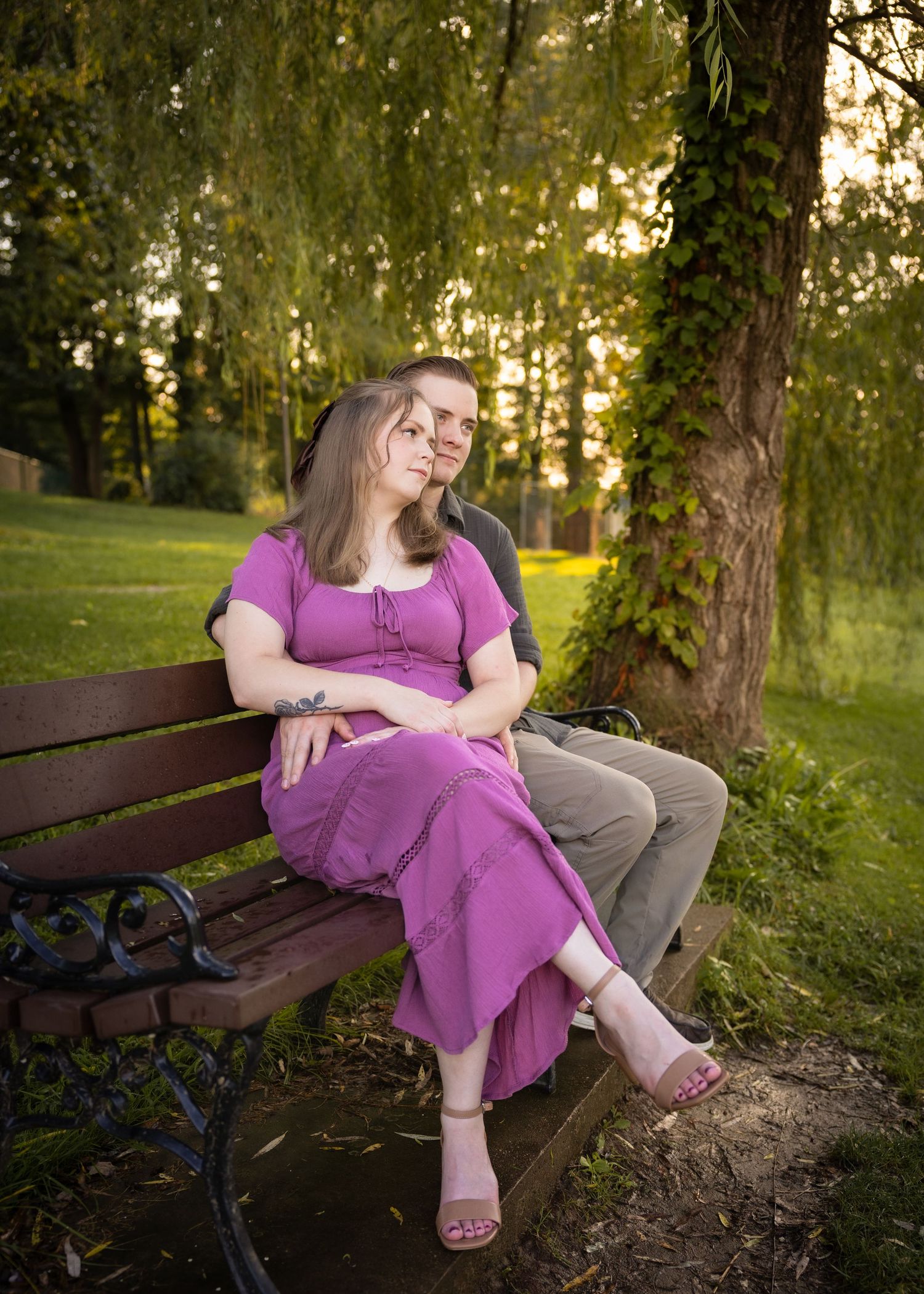 Couple sitting on a wooden park bench at golden hour, woman in purple dress leaning on man in gray shirt.