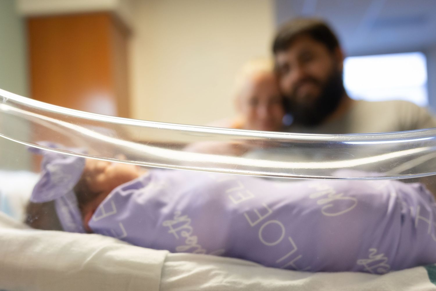 Newborn baby sleeping in hospital bassinet while father looks on lovingly in the background.
