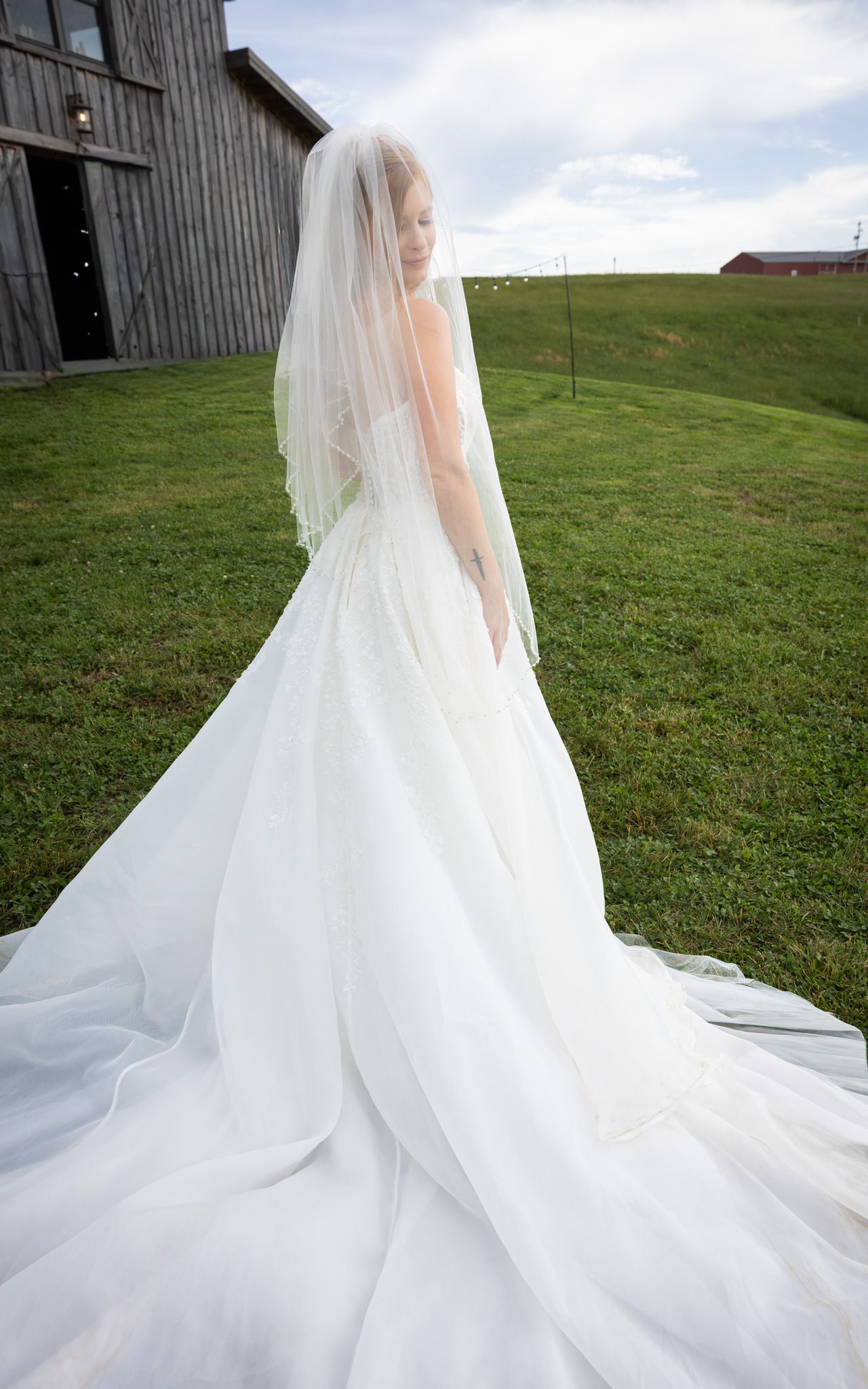 A bride in a flowing white gown and veil stands with her back to the camera near a rustic barn.