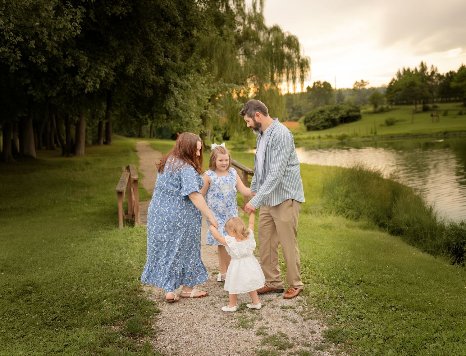 Family of four holding hands on a scenic path by a pond at golden hour, surrounded by lush green trees.