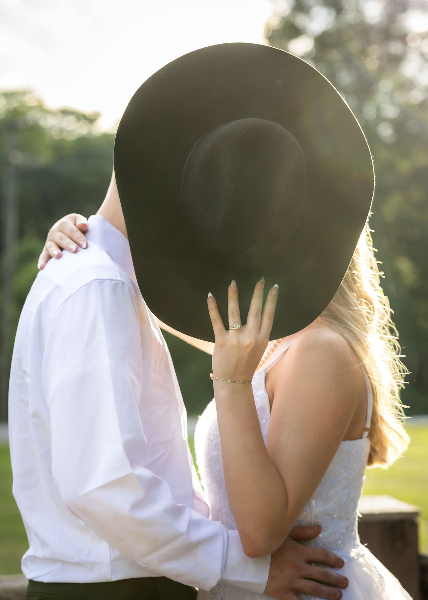 Couple shares a romantic kiss hidden behind a wide-brim black hat during outdoor wedding.