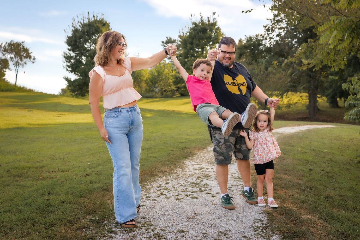 Family of four walking on a park path, parents swinging young child while toddler walks alongside on sunny day.