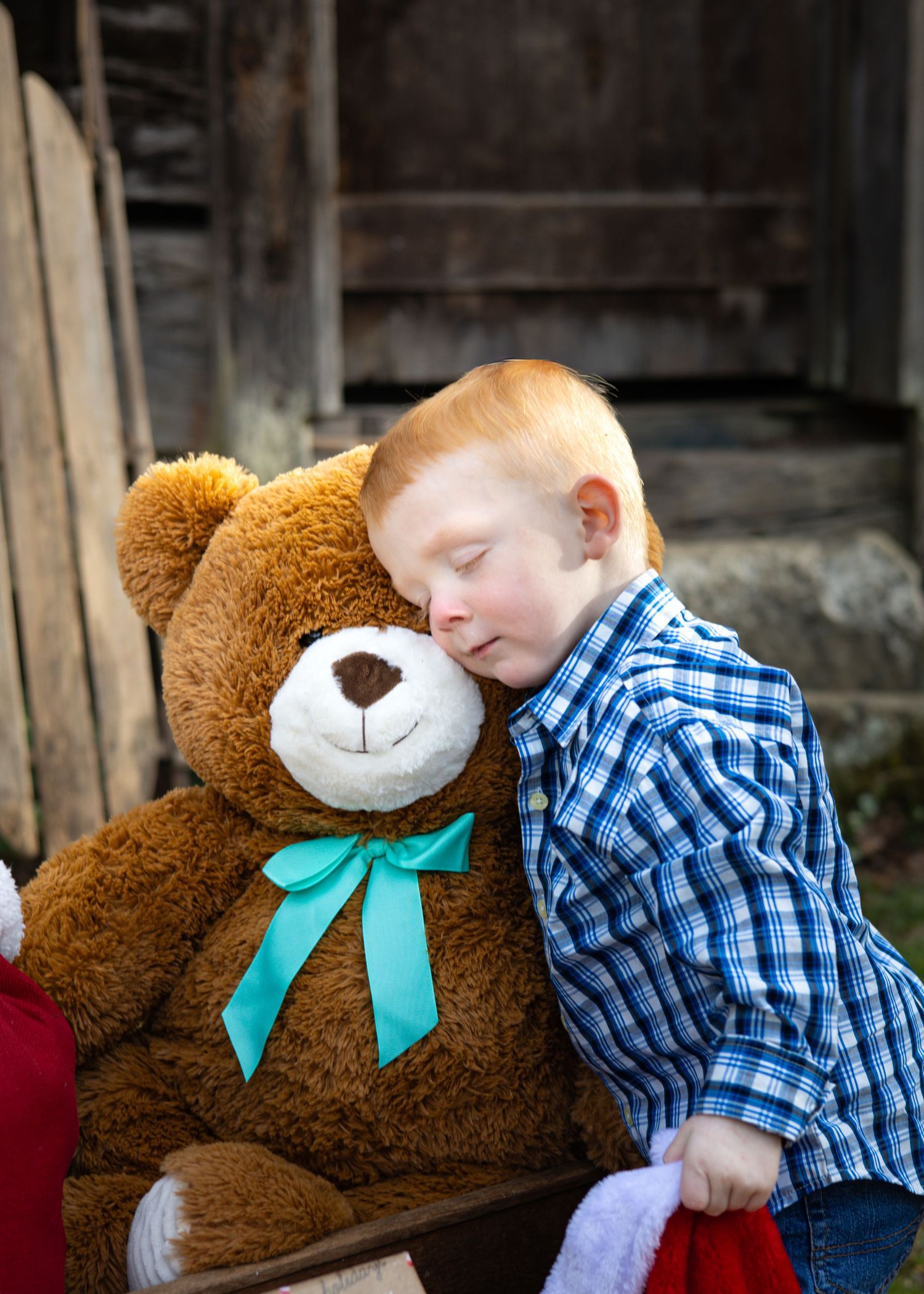 A red-haired boy rests his head on a large brown teddy bear with a teal bow outdoors.
