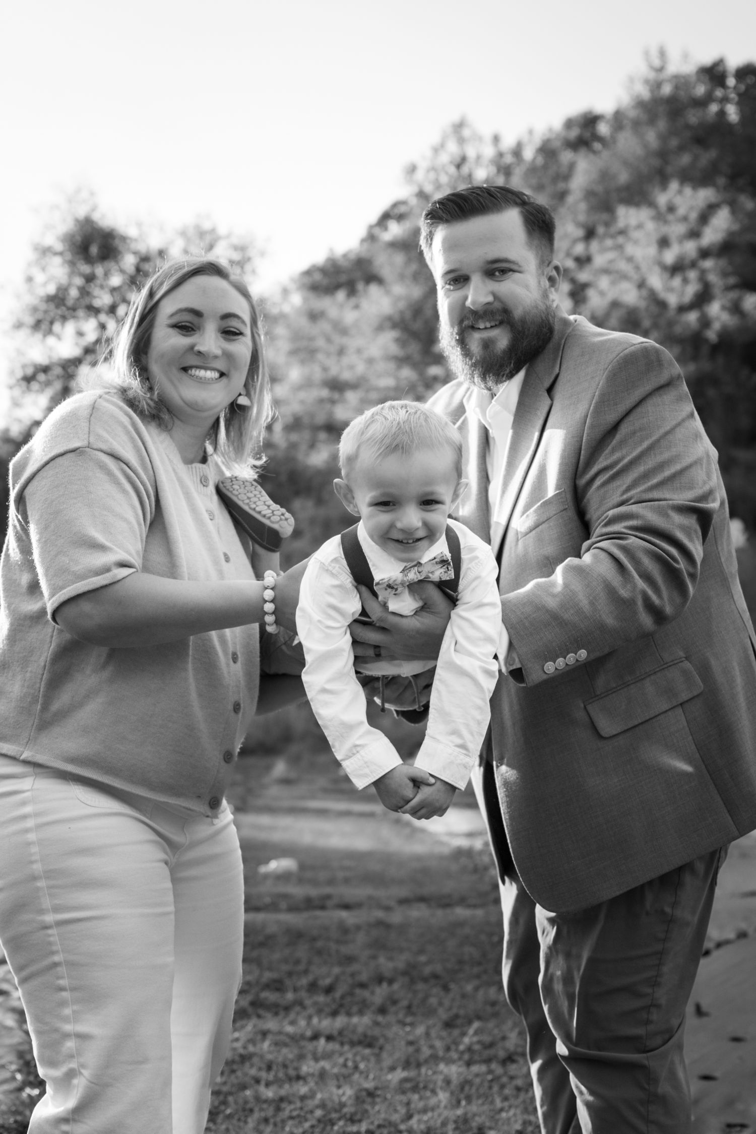 Black and white family portrait of smiling parents holding young boy in bow tie outdoors.