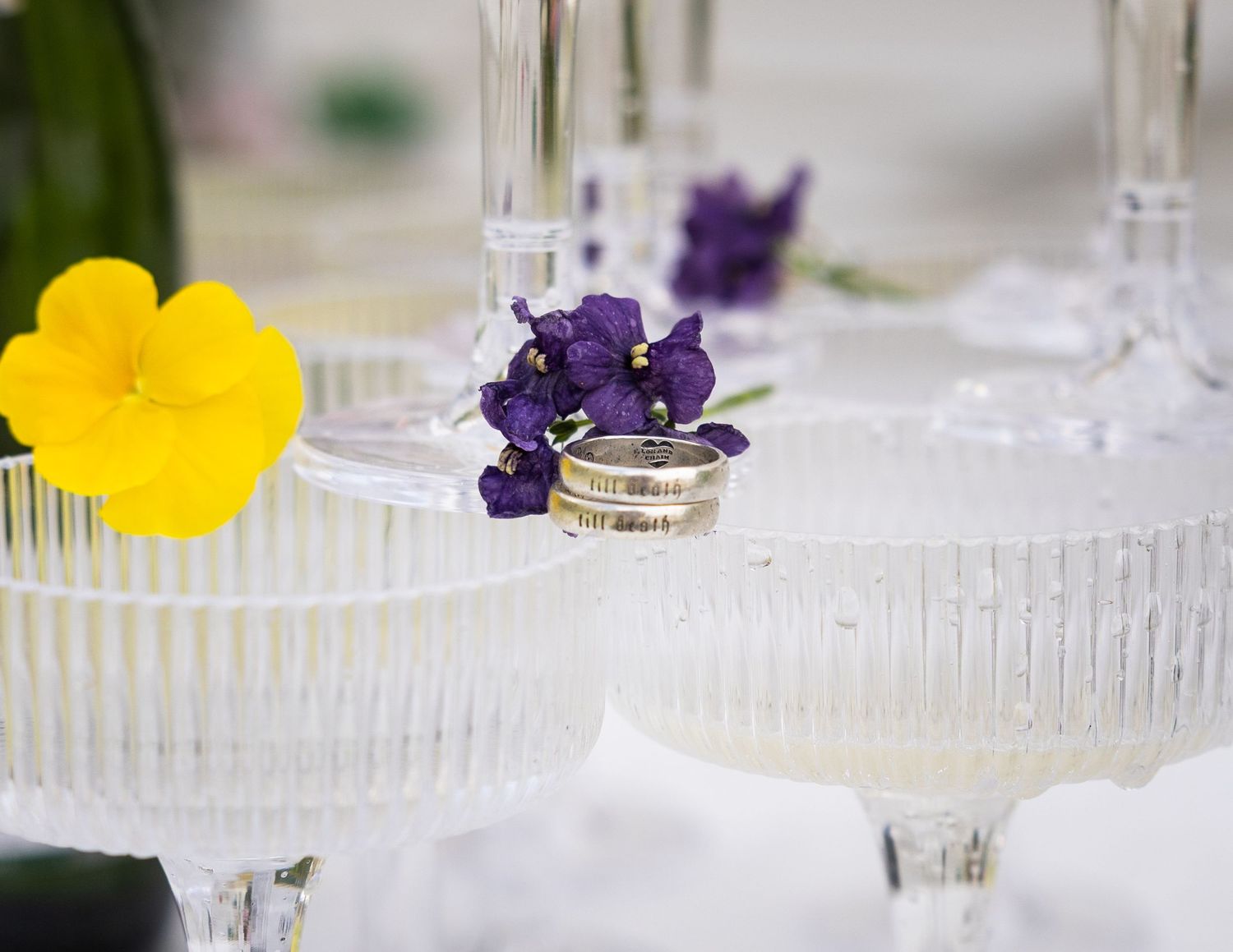 Wedding rings resting on champagne tower glasses with purple and yellow flowers.
