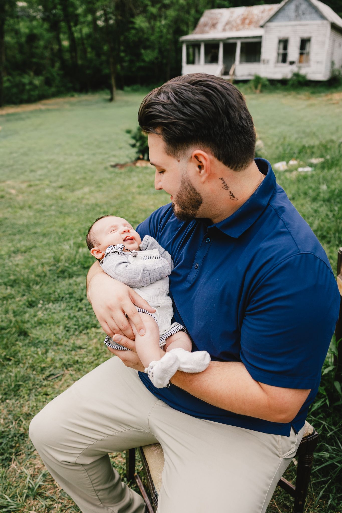 Proud father in blue polo shirt tenderly holding smiling newborn baby outdoors near rustic farmhouse.