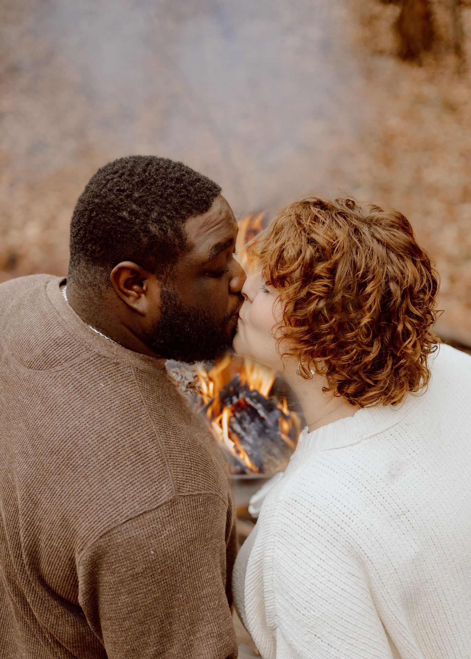 A couple shares a kiss in front of a glowing campfire surrounded by autumn foliage.