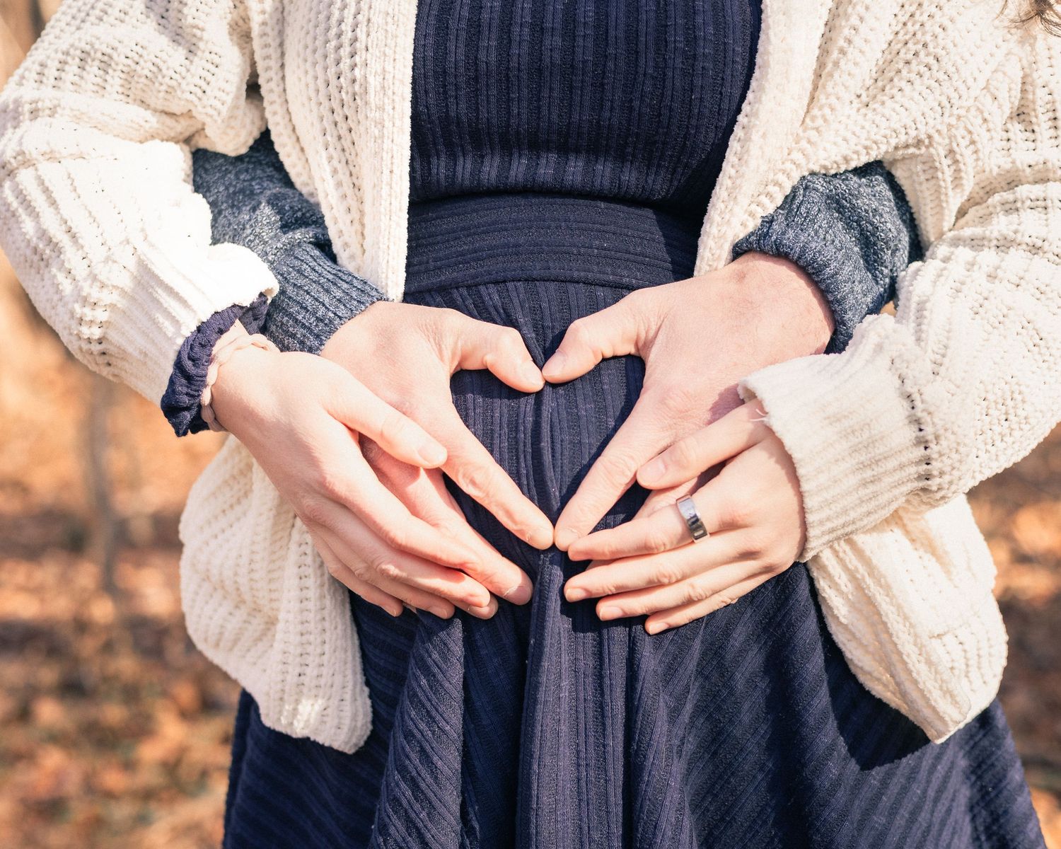 A couple forms a heart shape with their hands over a pregnant belly in an autumn outdoor setting.