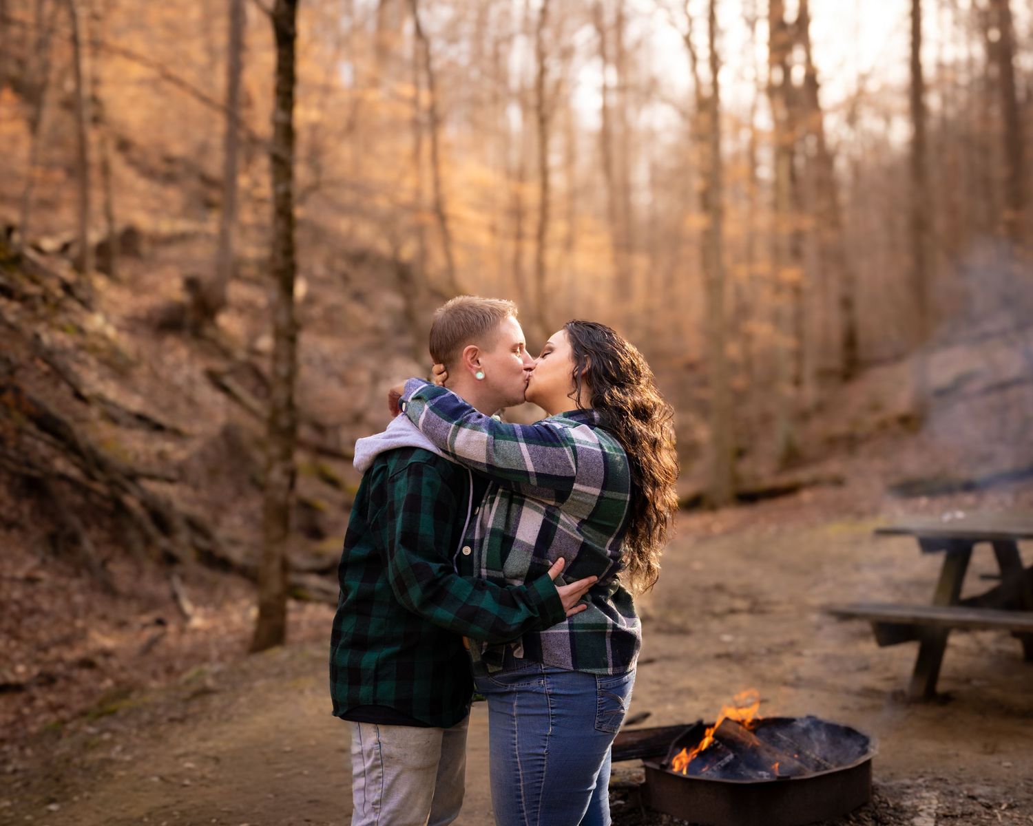 Couple kissing by a campfire in a golden autumn forest setting.