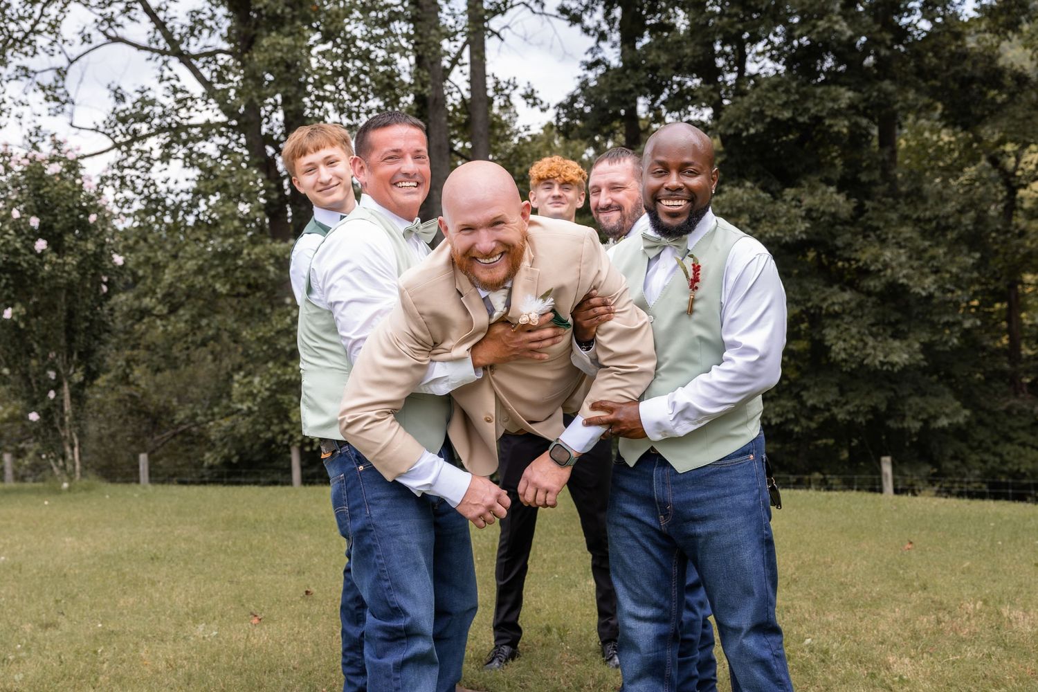 Groomsmen in sage vests and jeans laughing and playfully lifting the groom outdoors at a wedding.