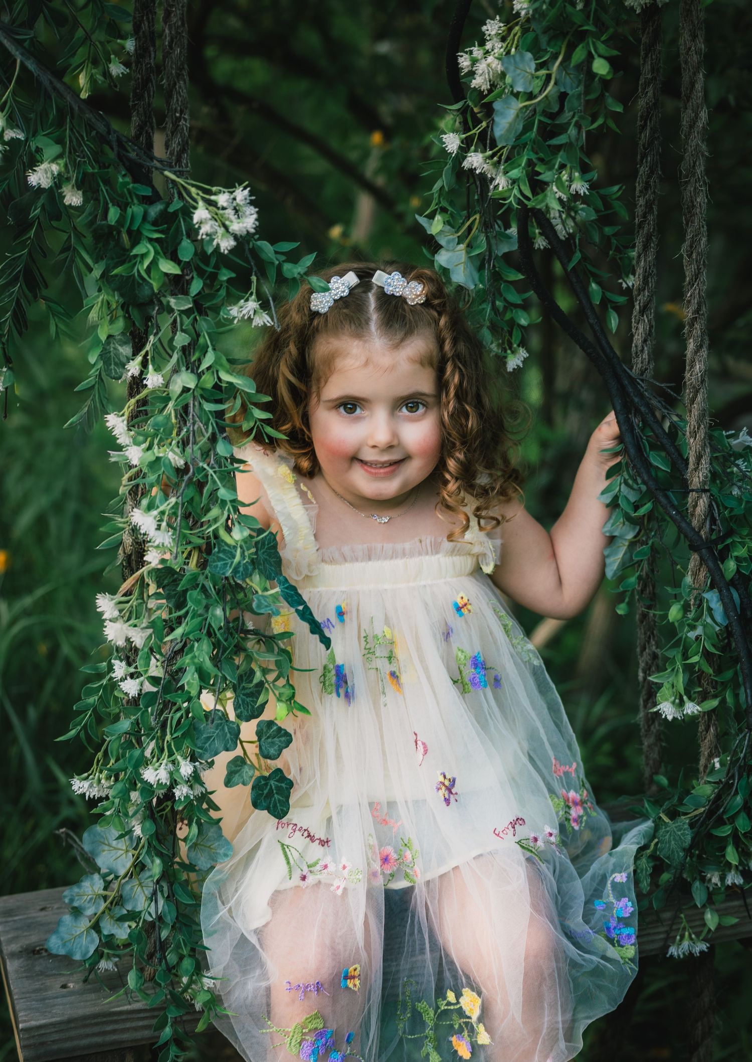 Adorable girl in floral tulle dress sitting on vine-covered swing in garden.