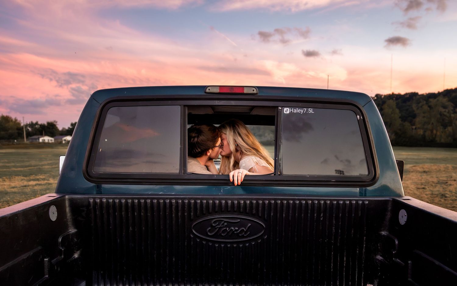 Couple sharing a romantic kiss in the back window of a Ford pickup truck at sunset with a colorful pink sky.