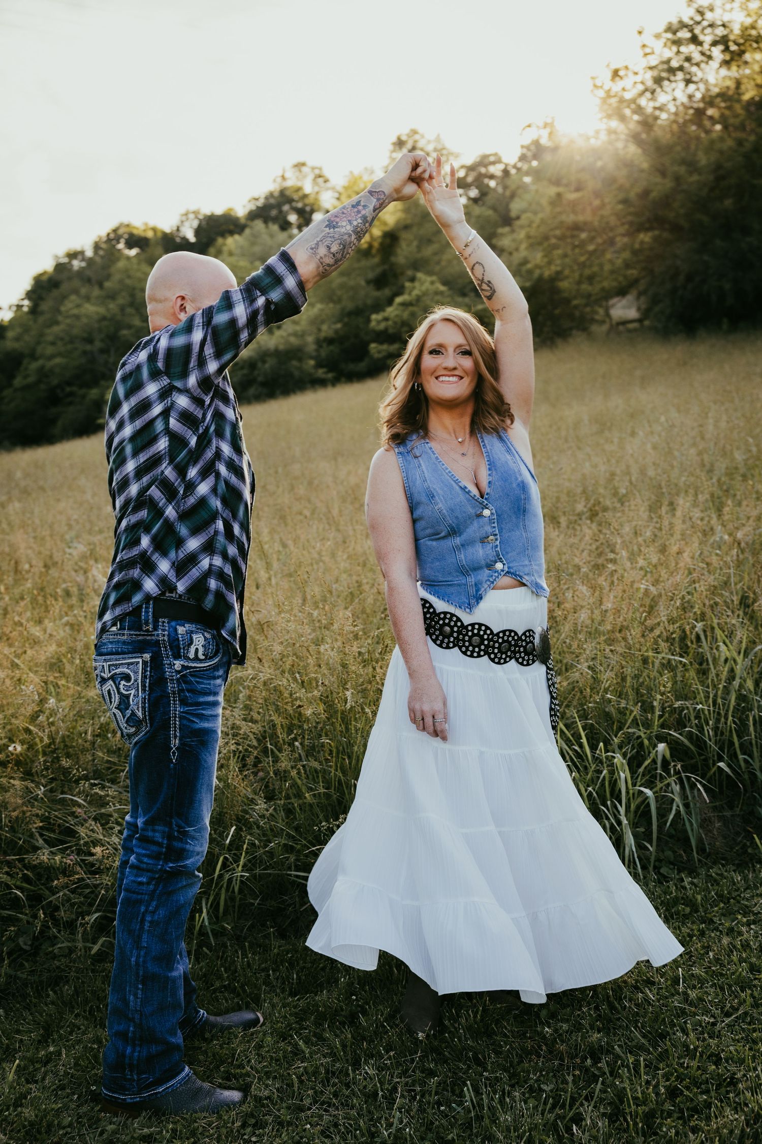 Couple dancing in a sunny field, man in plaid shirt twirling smiling woman in white skirt and denim vest.