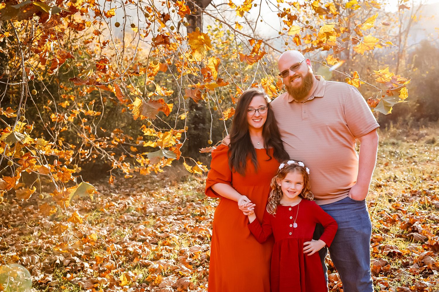 Family of three posing among vibrant autumn foliage, wearing red and pink outfits in a warm golden fall setting.