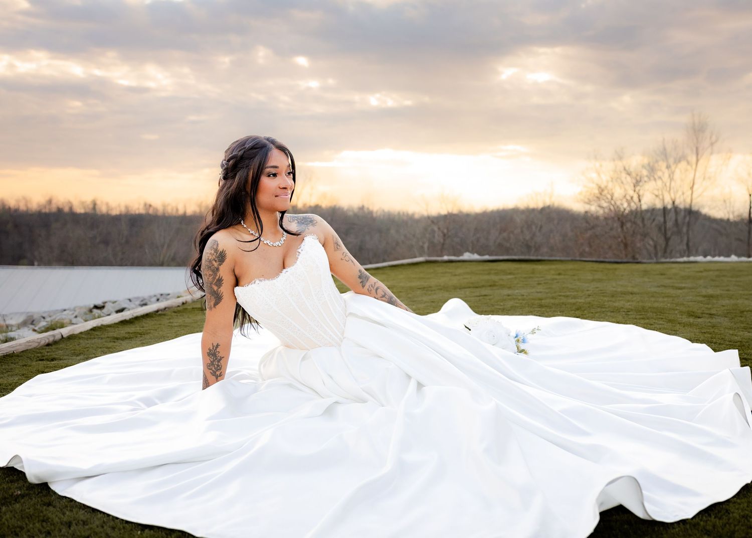 Bride in a stunning white ball gown with long train sits on green grass at sunset near a serene lake.