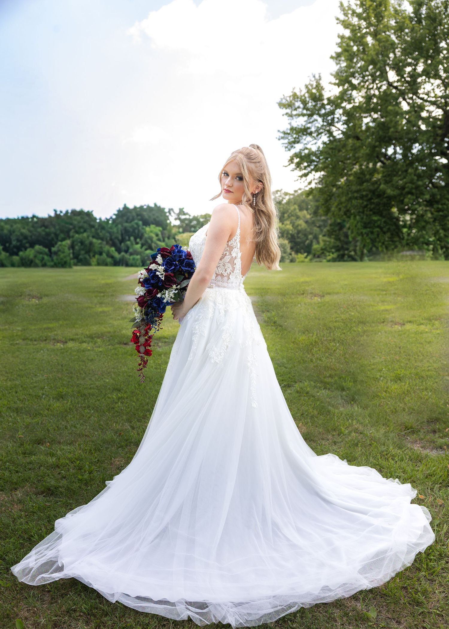 Bride in white lace wedding gown holding blue and red floral bouquet, standing in green field with trees.