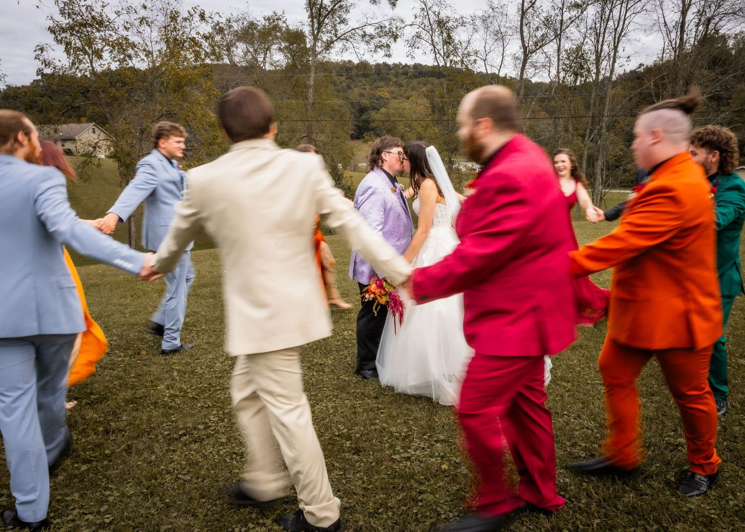Wedding party in colorful suits dancing in a circle outdoors on green grass with trees in background.