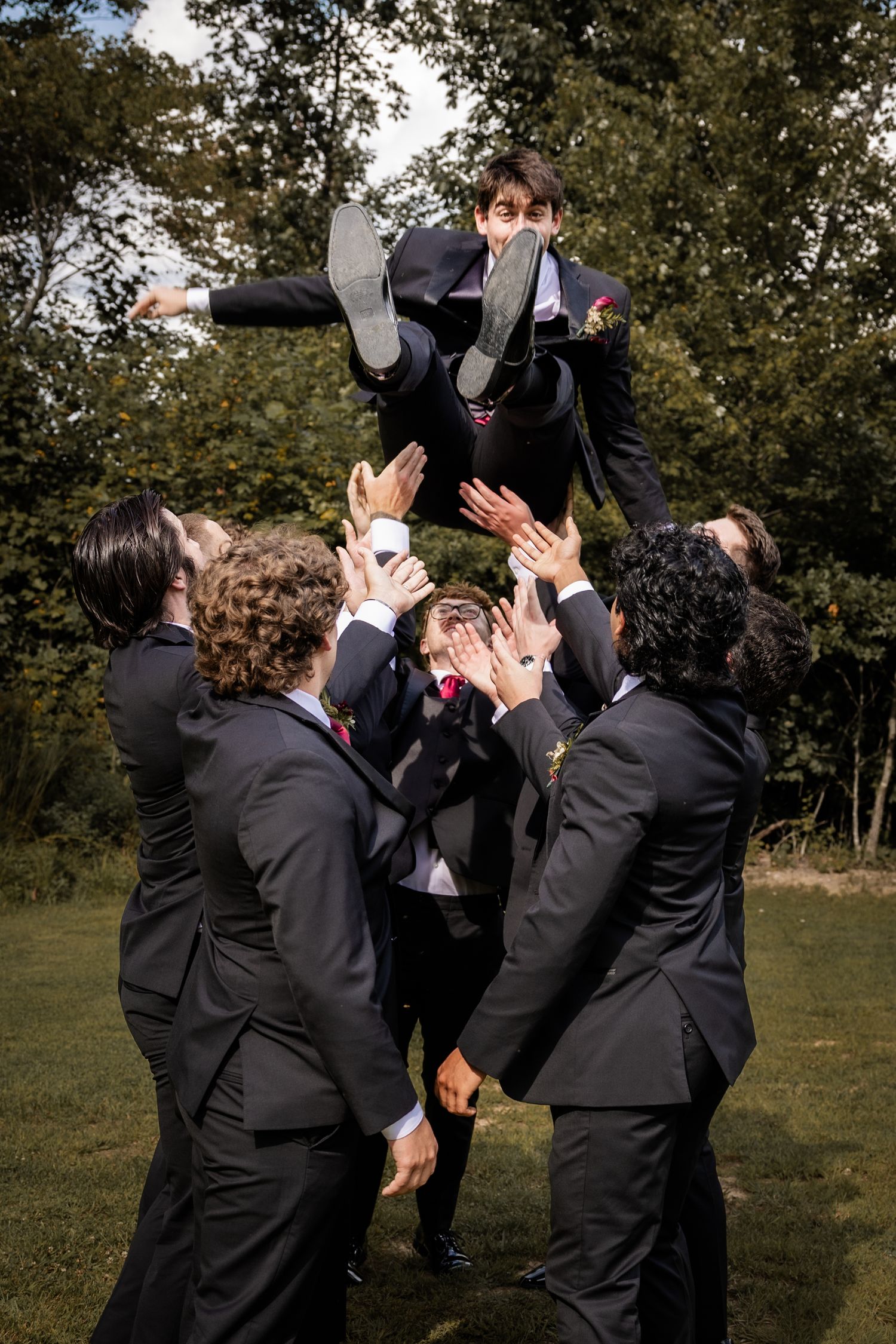 Groomsmen in black suits tossing the groom into the air outdoors, celebrating at a wedding reception.