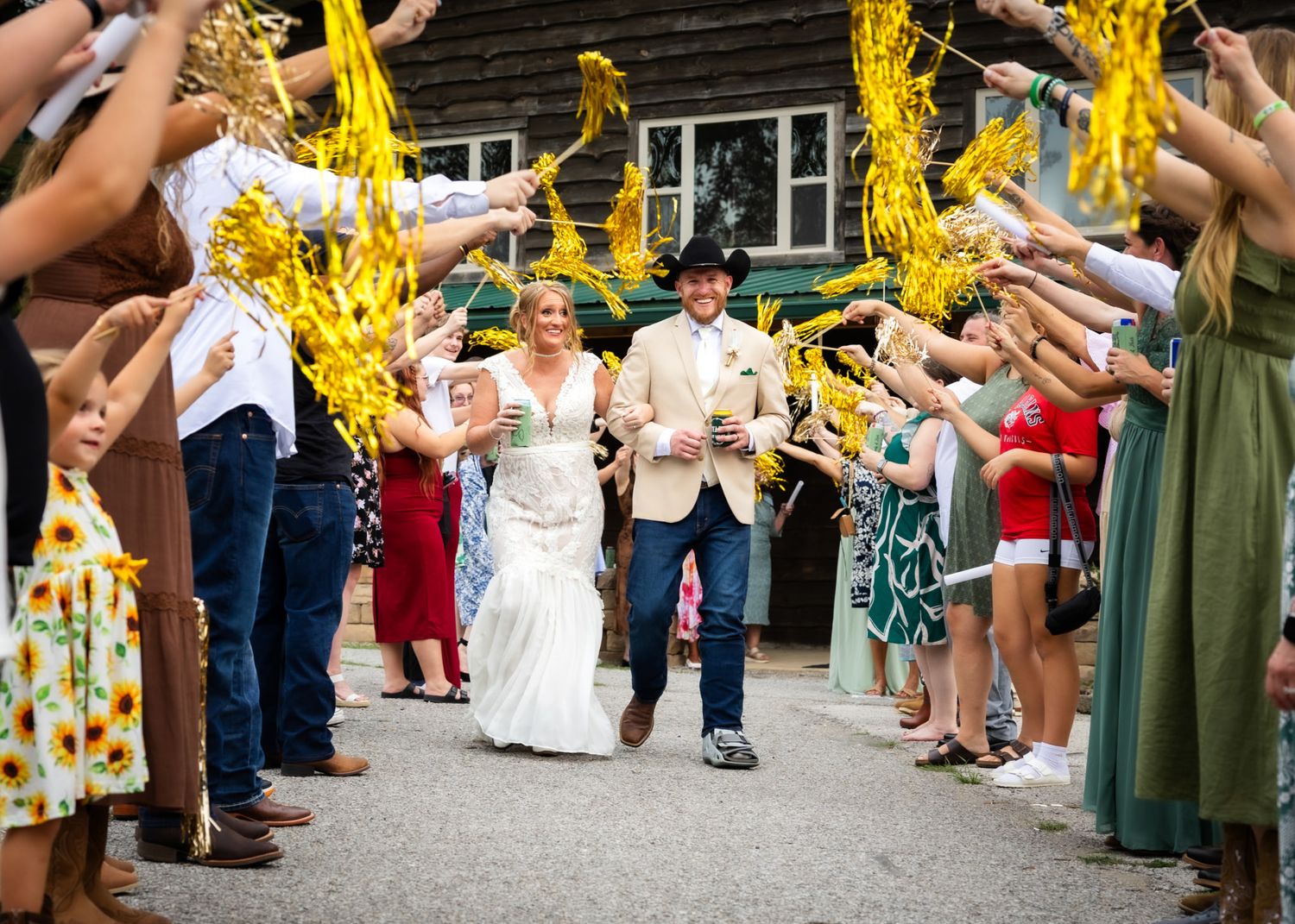 Bride and groom walk through a joyful wedding sendoff tunnel of guests holding yellow ribbon wands outdoors.
