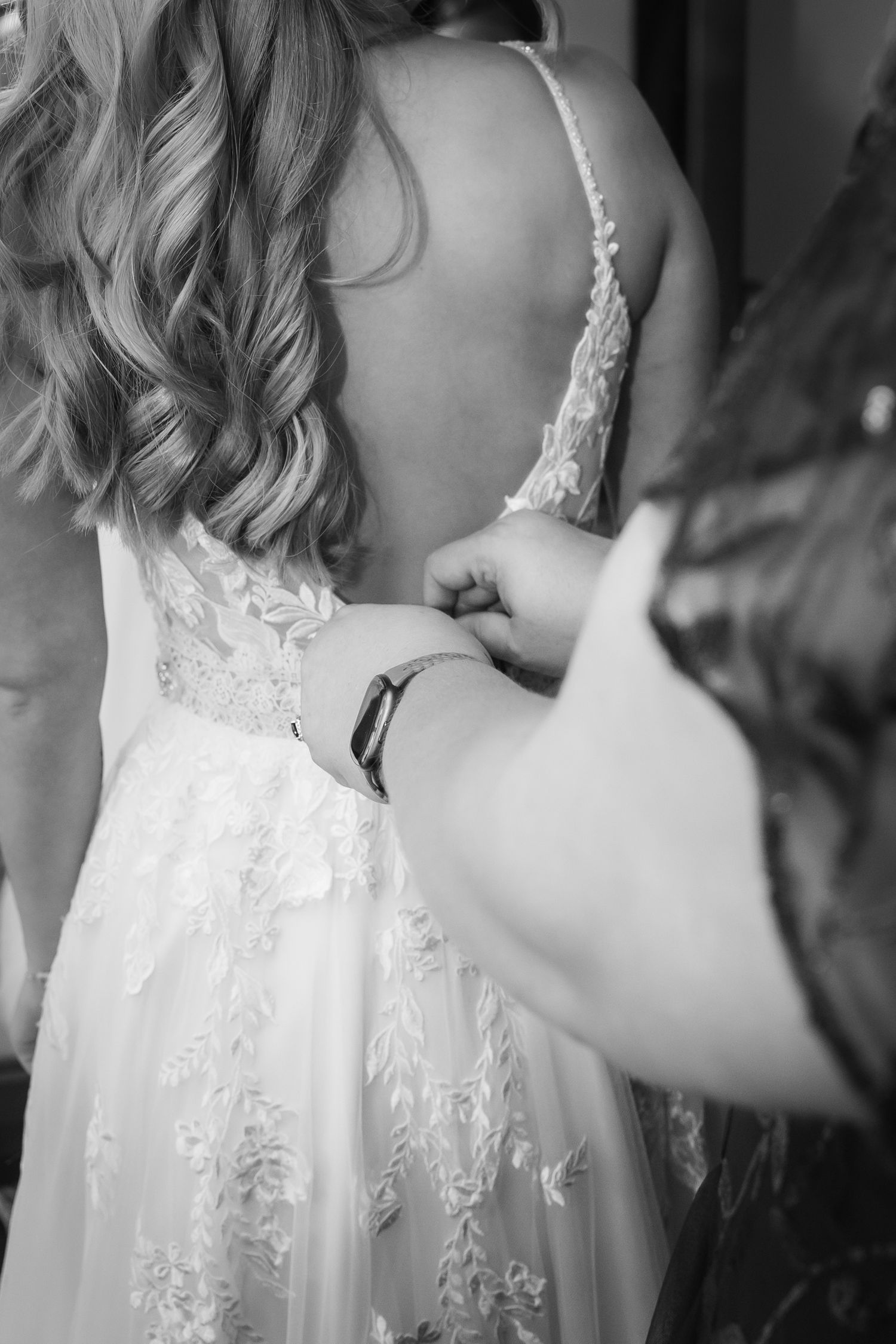 Black and white photo of a bride in lace wedding dress being buttoned up by a bridesmaid.