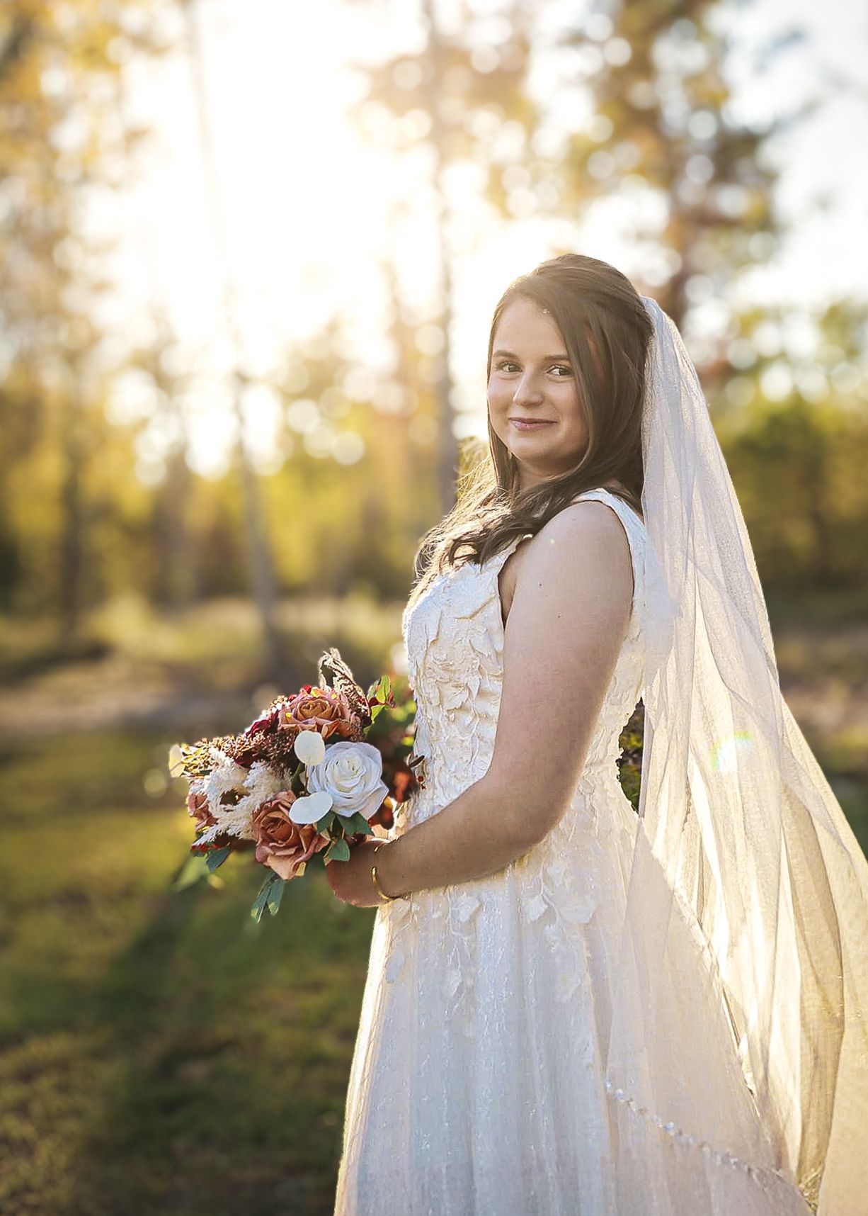 Bride in white lace gown holds floral bouquet in golden sunlit outdoor setting.