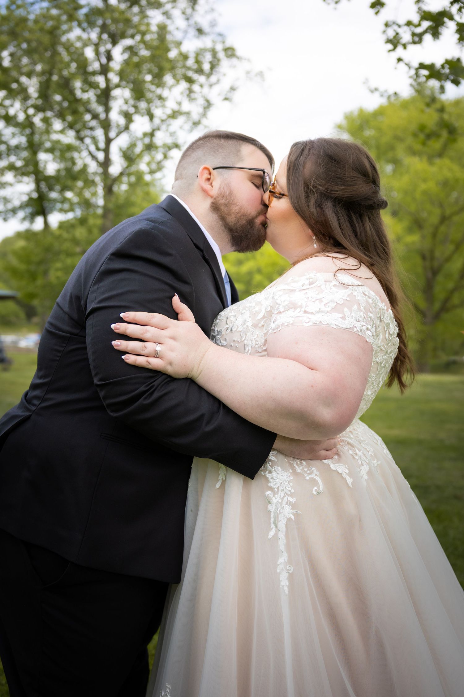 A bride and groom share a romantic kiss outdoors surrounded by lush green trees on their wedding day.