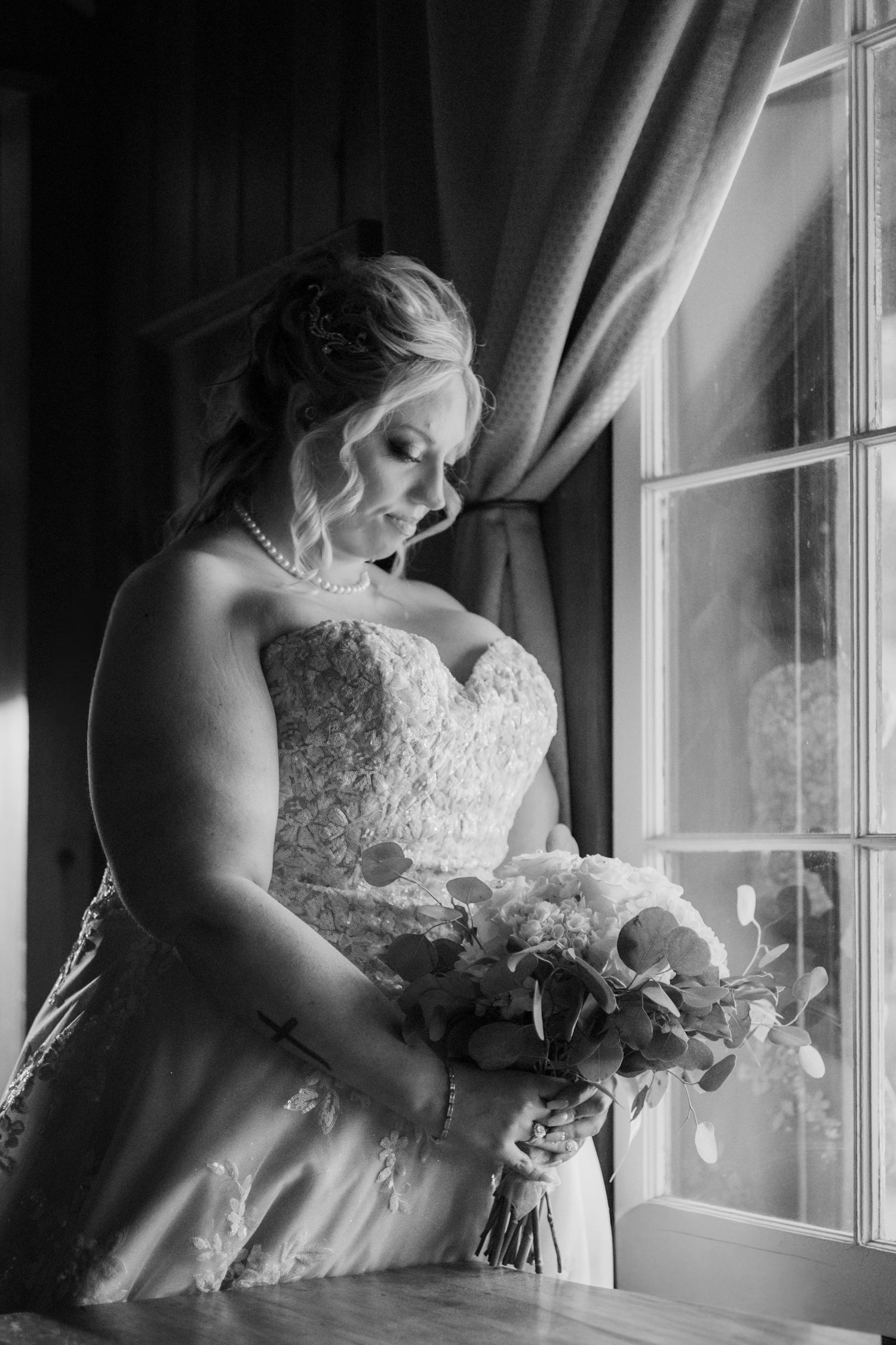 Black and white photo of a bride in lace gown holding a bouquet, gazing down by a sunlit window.