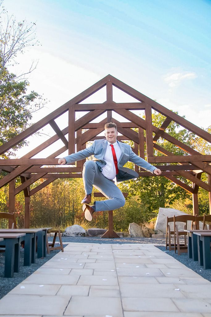 Groom in gray suit jumping joyfully under a rustic wooden outdoor wedding ceremony structure.