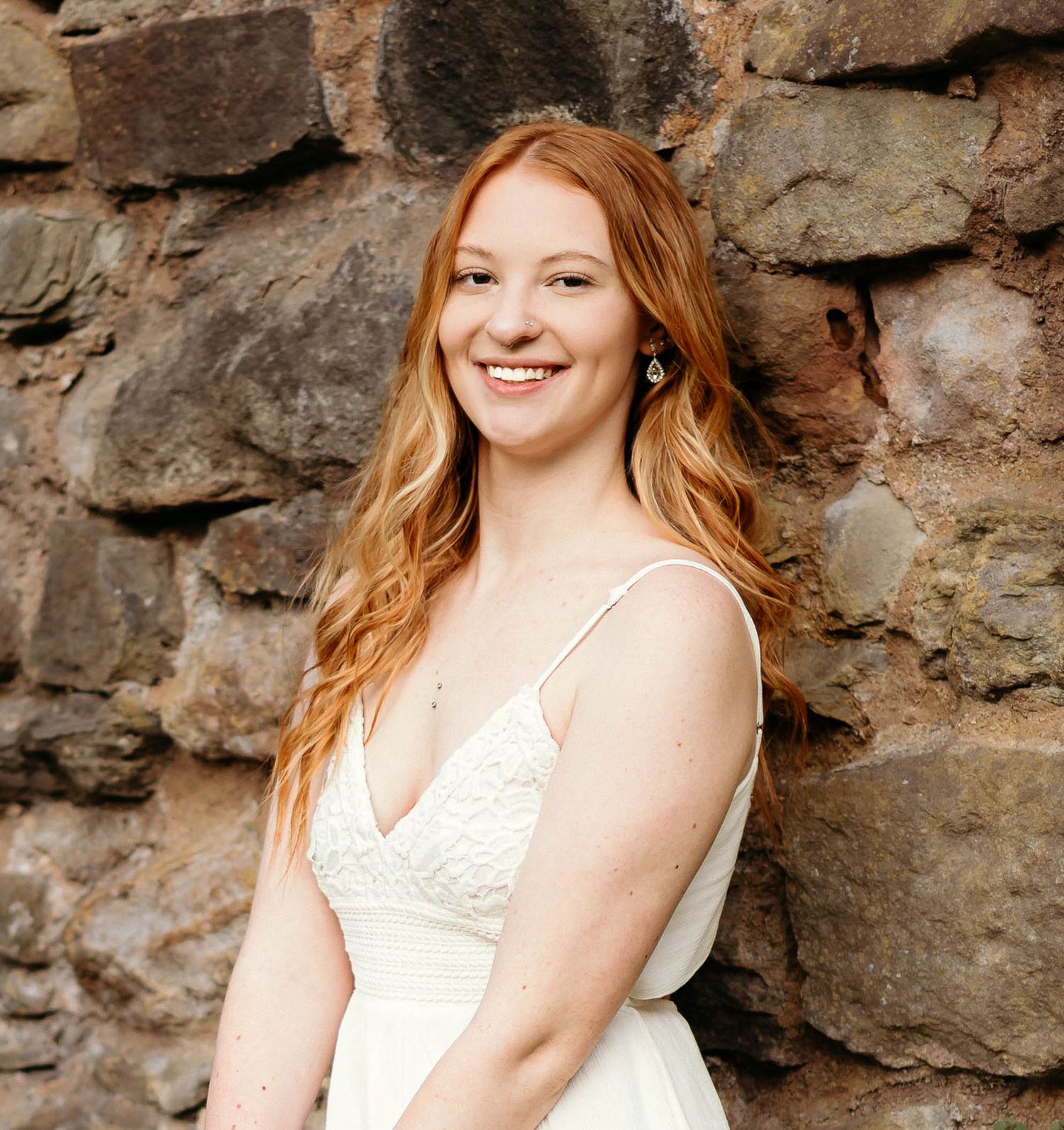 Smiling young redhead woman in white lace dress leaning against rustic stone wall.