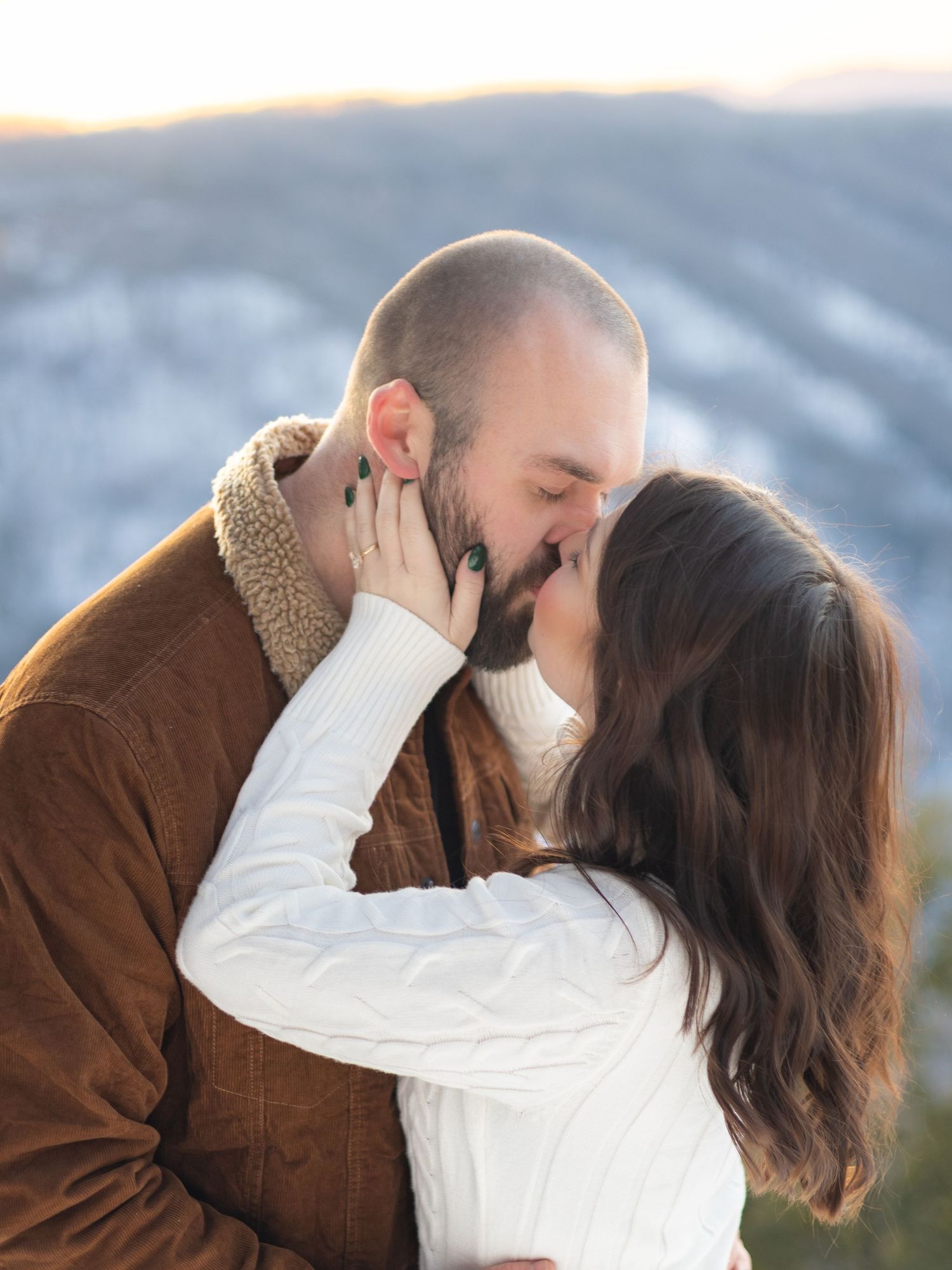 Couple sharing romantic kiss at snowy mountain overlook with soft winter light background.