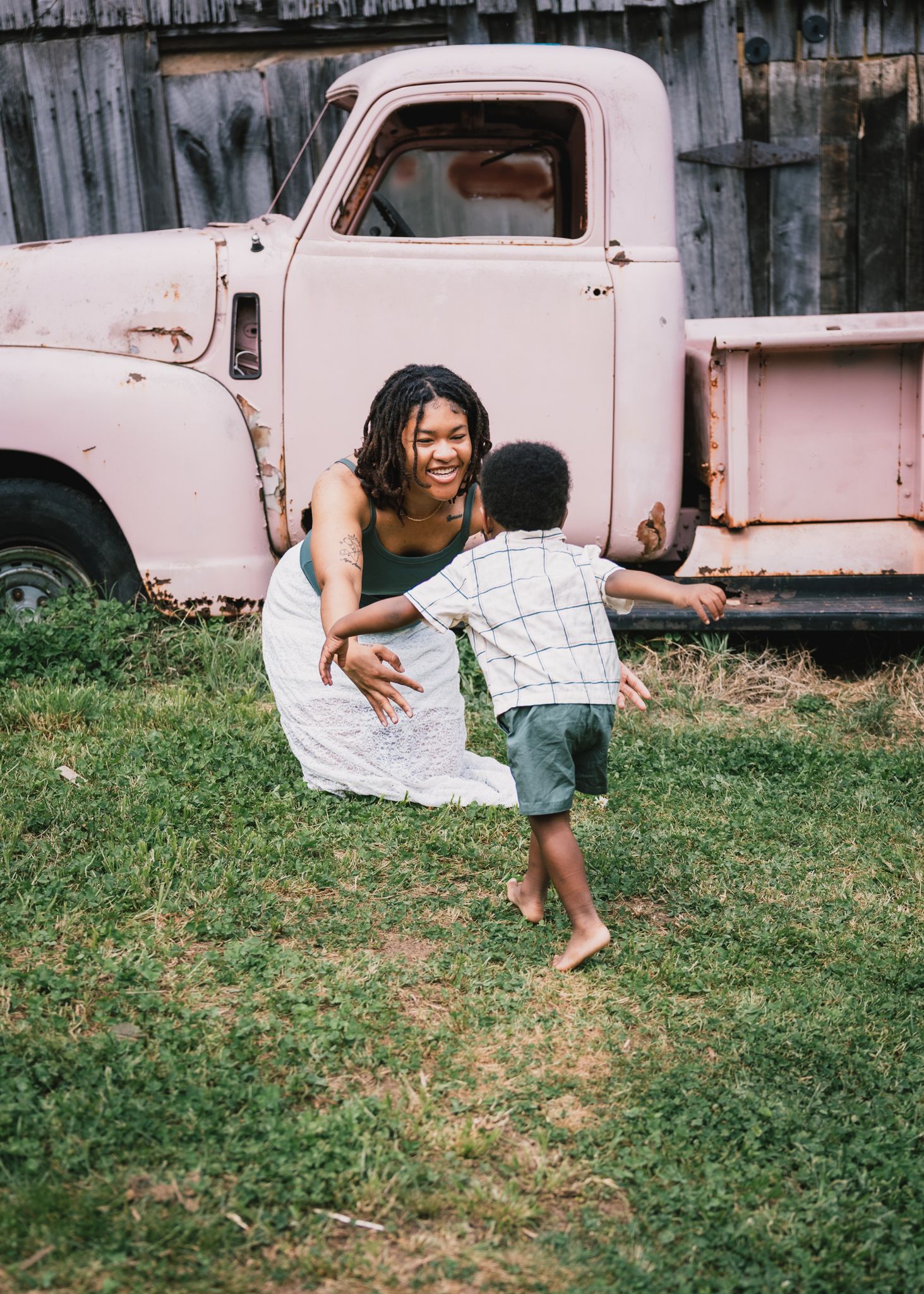 A joyful Black mother opens her arms to welcome her running toddler son near a vintage pink truck.