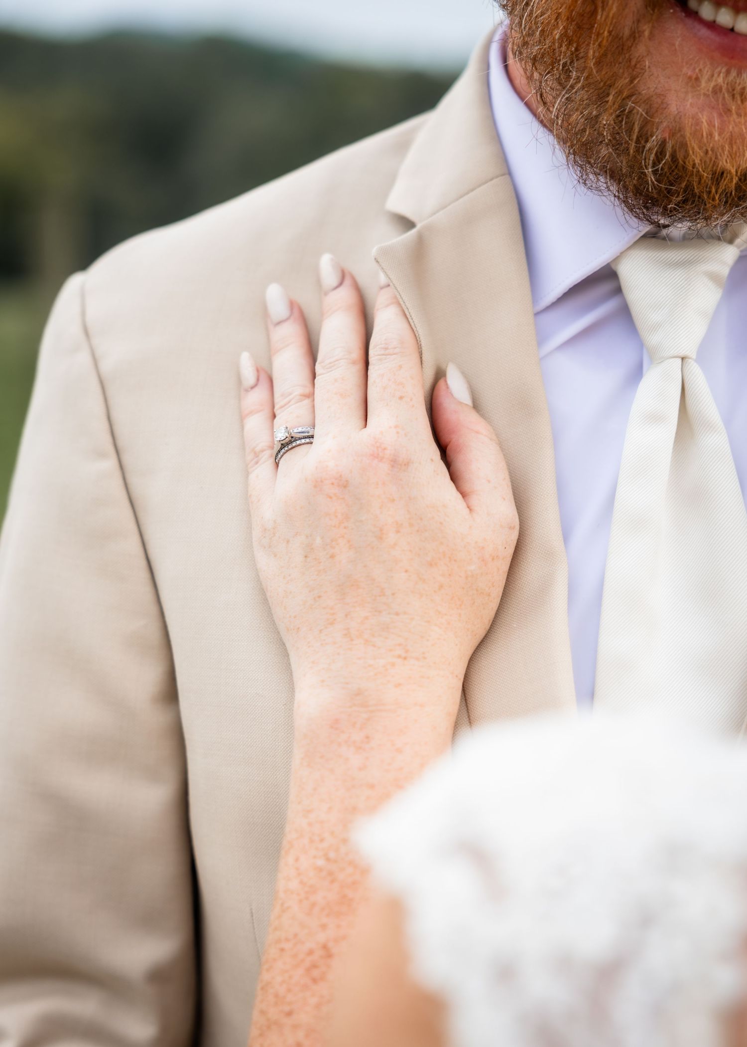 Bride placing hand on groom's tan suit chest, showing wedding ring, at outdoor ceremony.
