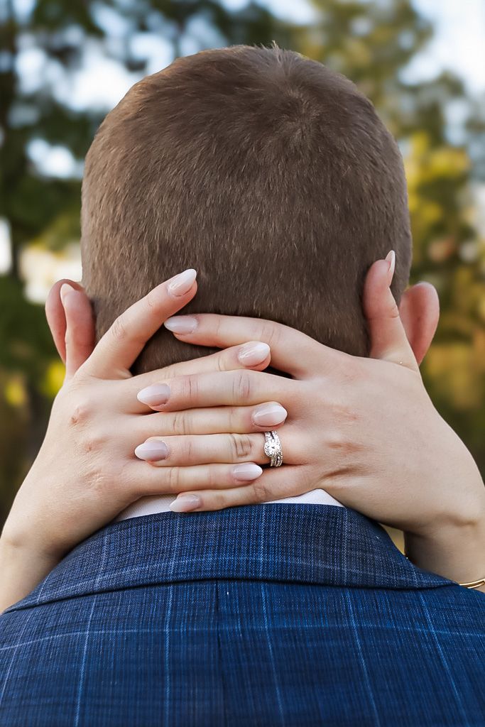 Close-up of bride's hands with diamond ring embracing groom's neck outdoors.