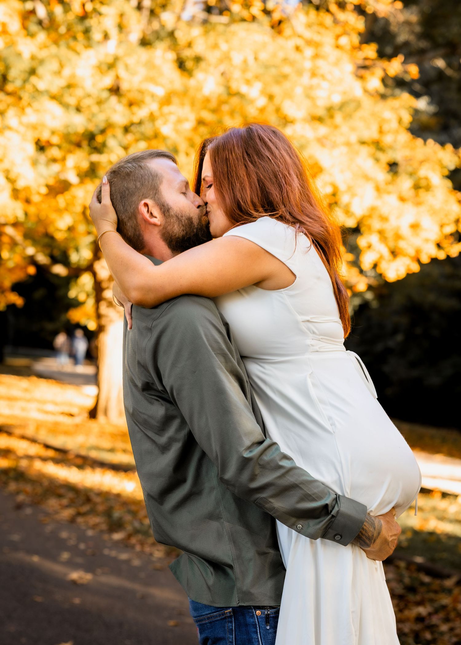 Expecting couple in a white dress and olive shirt share a kiss outdoors beneath golden autumn trees during a maternity photo session.