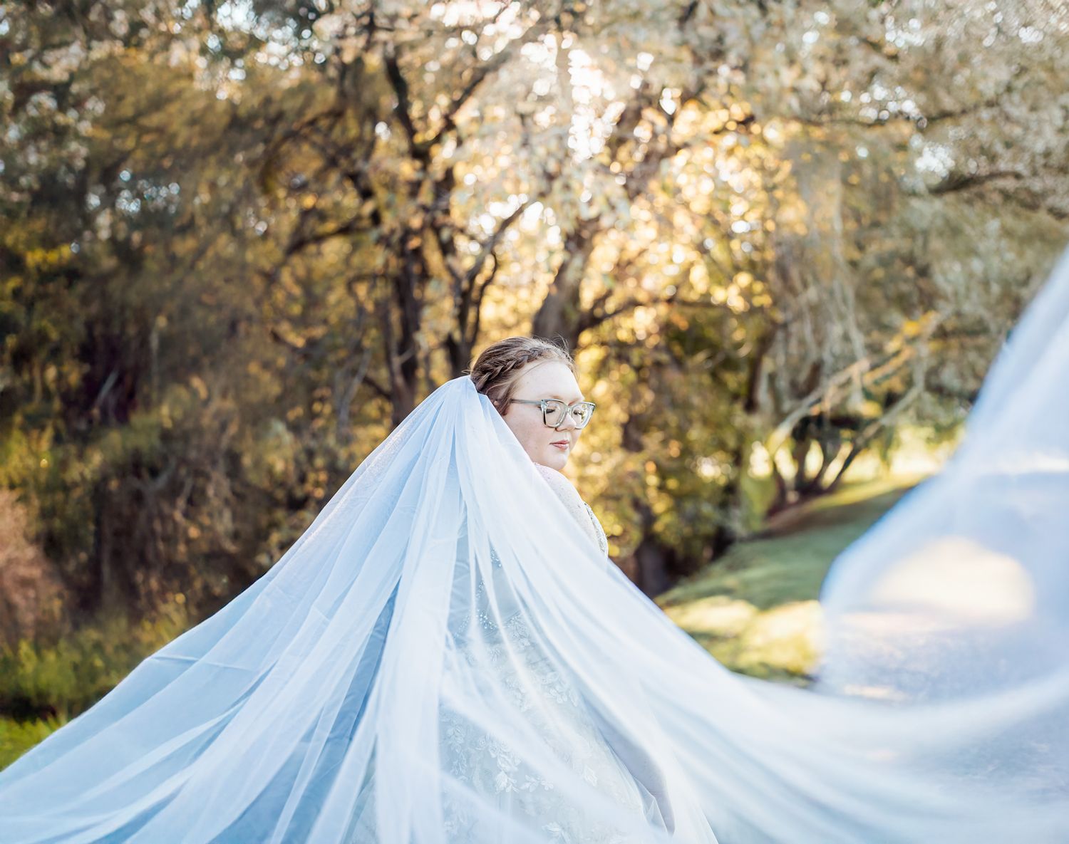 Bride in white gown with flowing veil billowing in sunlit outdoor garden setting.